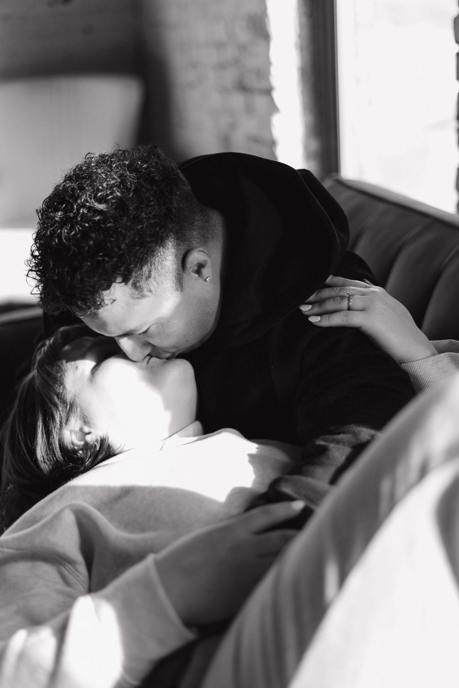 Black and white photo of a couple sharing a tender kiss on a couch, with soft natural light streaming in.