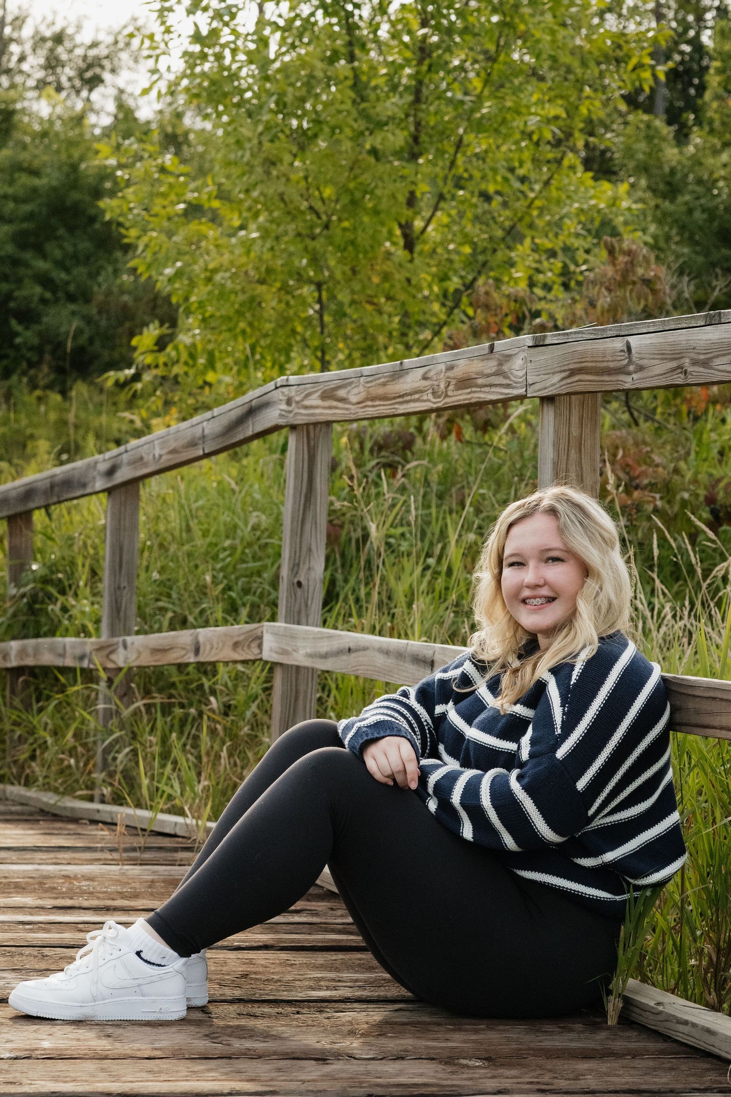 Smiling blonde teen in striped sweater sits on wooden bridge surrounded by autumn greenery.