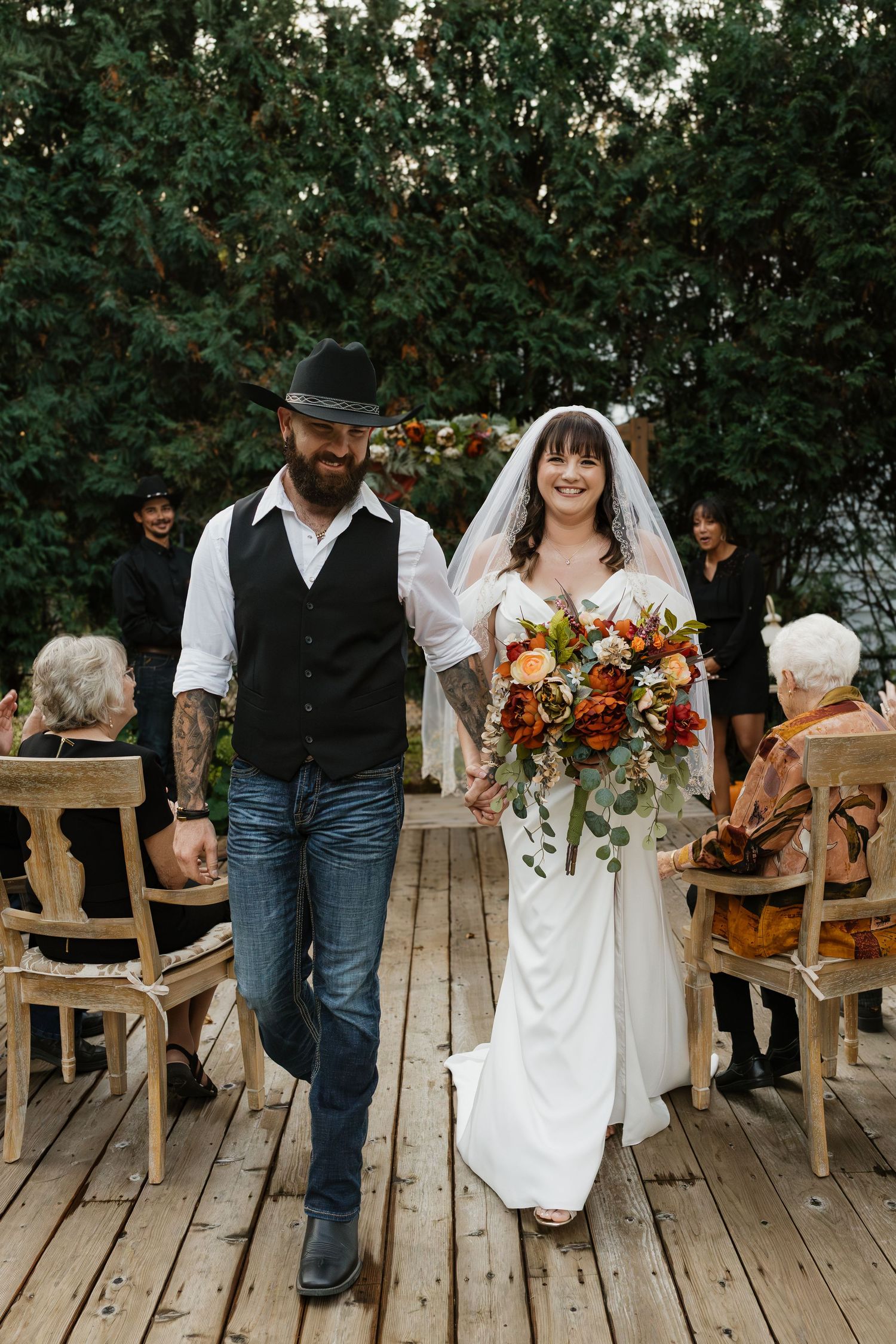 Smiling bride and groom walk down aisle after outdoor fall wedding ceremony on rustic wooden deck.