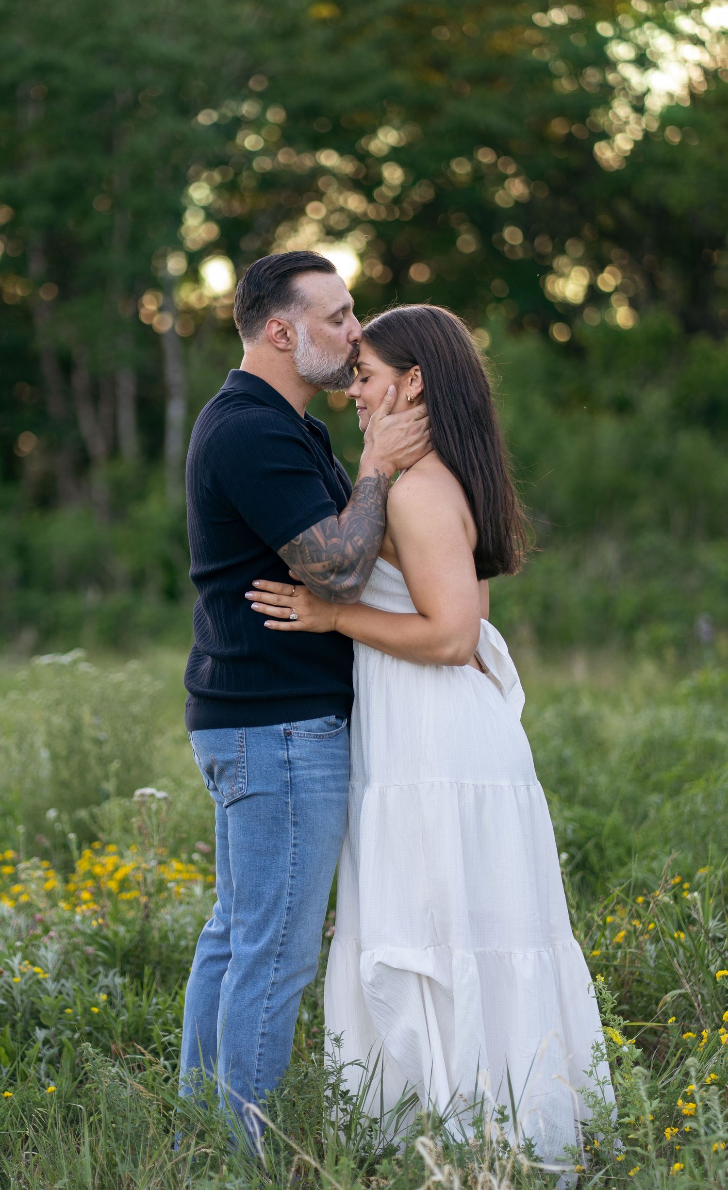 A couple shares a romantic moment in a wildflower field, man in black shirt and jeans kissing woman in white dress.