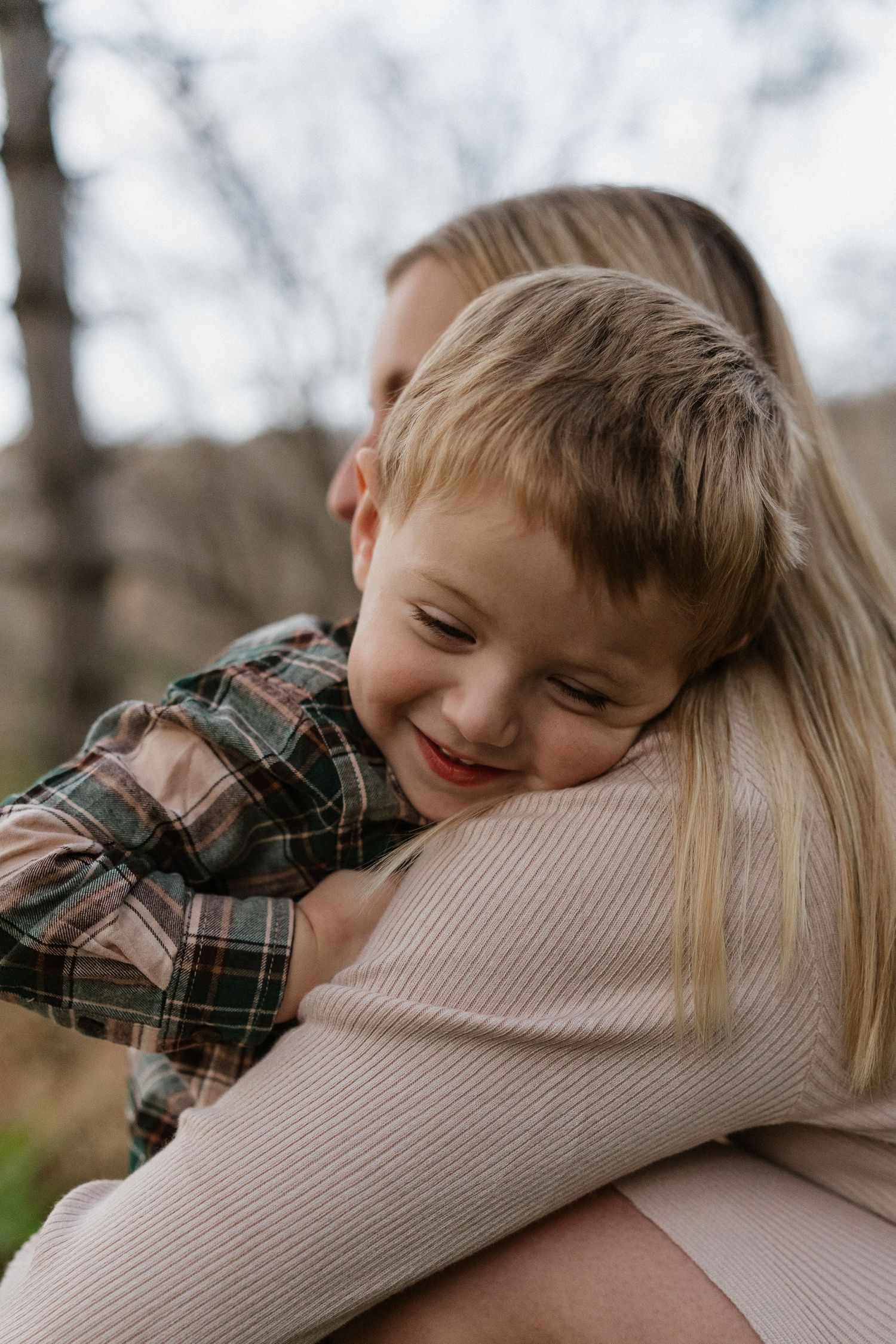 Young boy in plaid shirt smiles while resting his head on mothers shoulder during outdoor photo session.