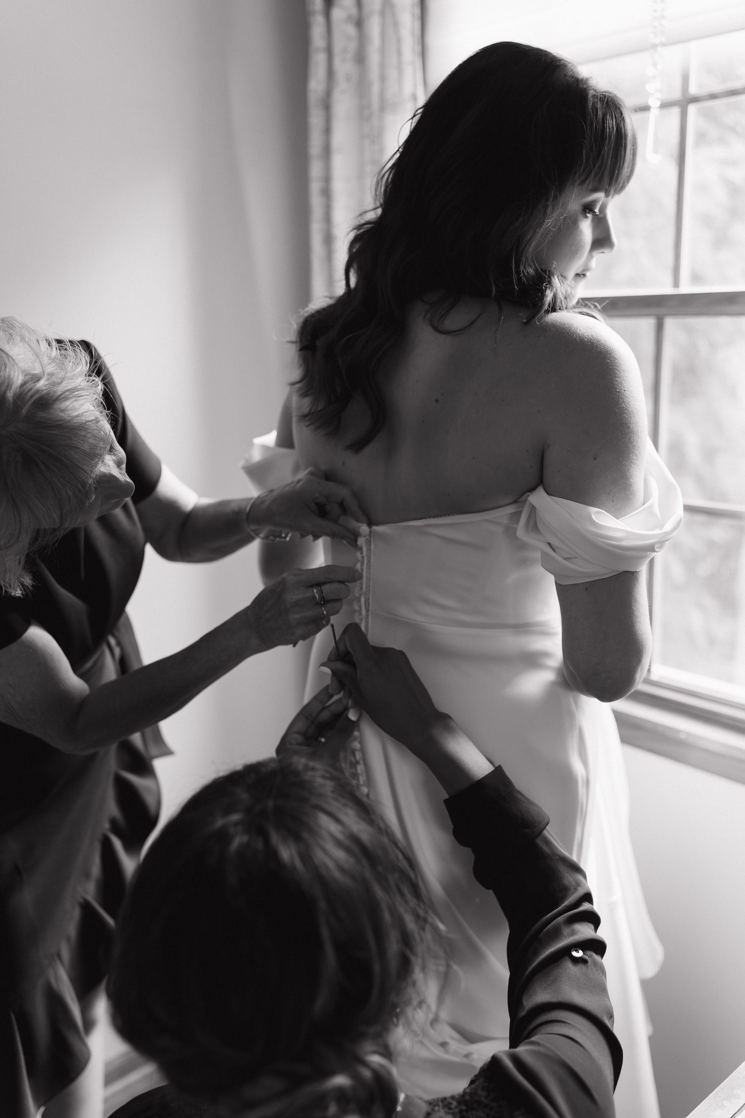 Black and white photo of bride being helped into wedding dress by two people near window.