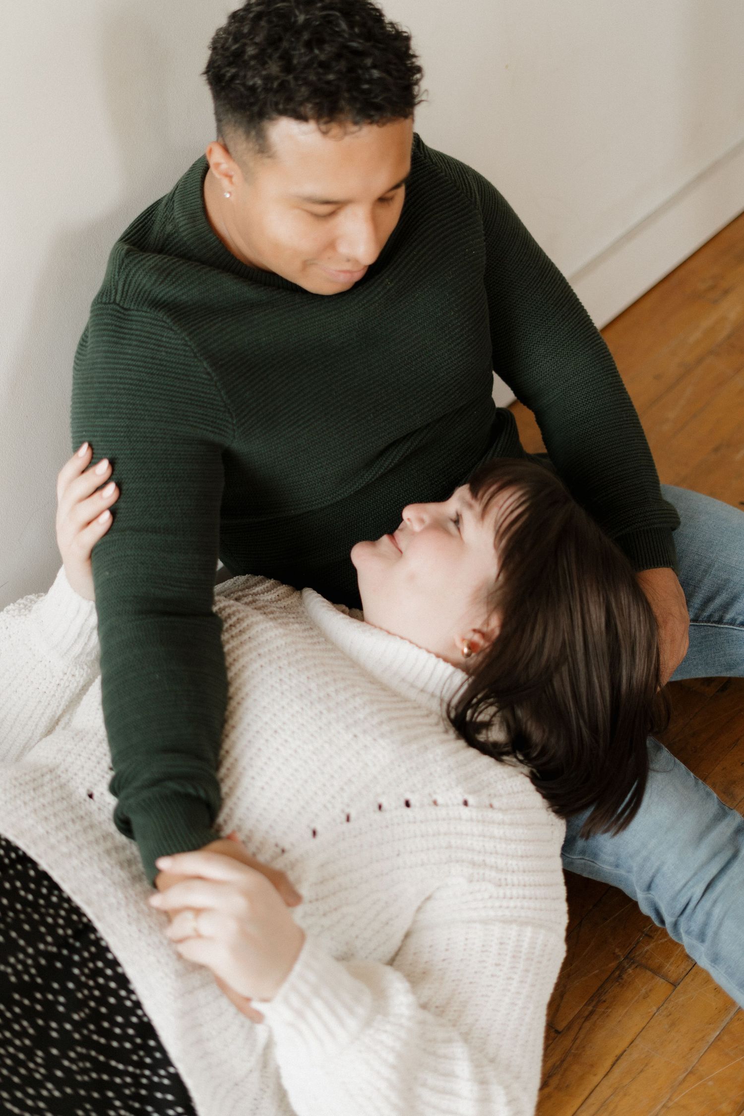 A couple shares a tender moment, woman lying on floor in white sweater, man in green sweater gazing down lovingly.