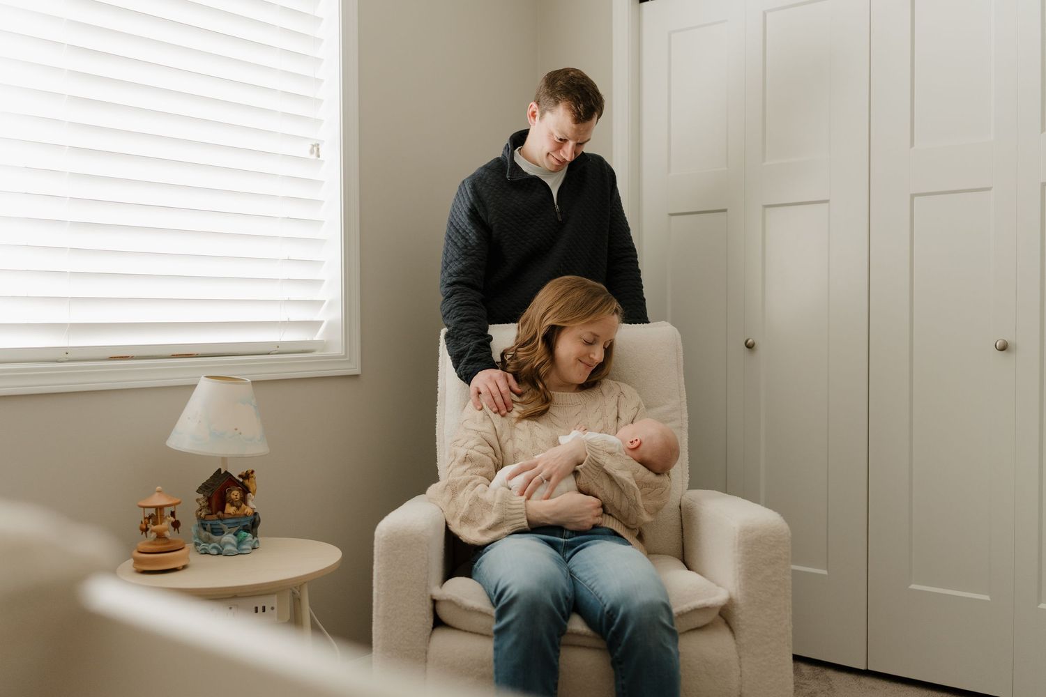 A couple sits in a nursery chair, tenderly holding their newborn baby in a softly lit room.