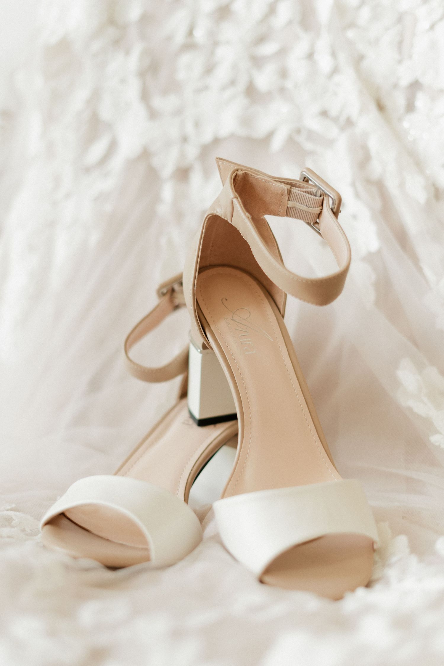 Elegant white bridal heels with ankle strap and block heel displayed against a white lace wedding dress.