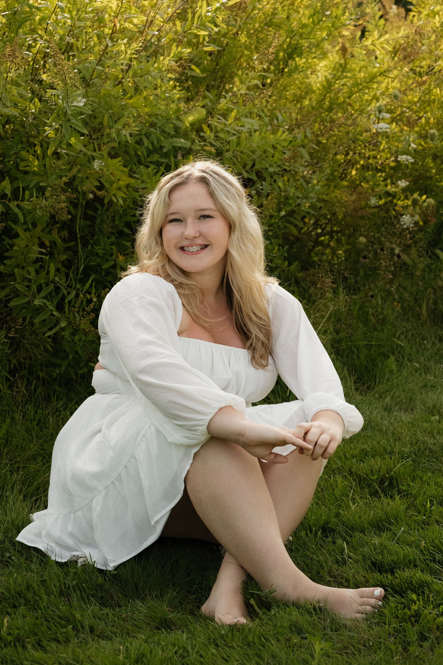 Smiling blonde teen in white dress sitting barefoot on green grass with lush foliage in the background.