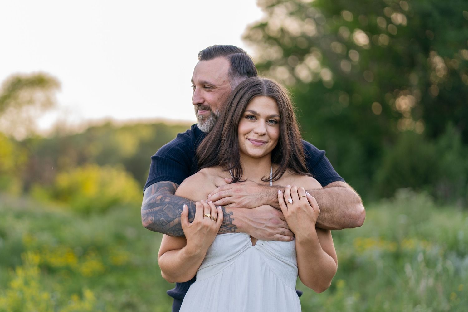 Couple embracing outdoors in a green field, man with tattoos hugging smiling woman in white dress at golden hour.