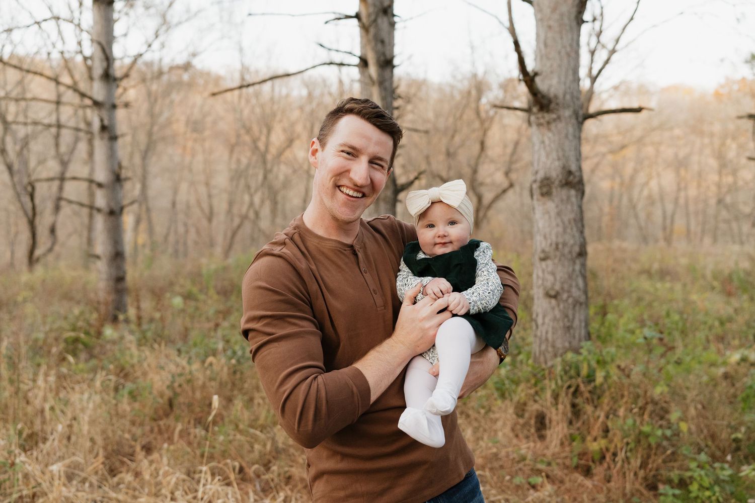 Smiling man in brown sweater holds baby girl in white bow headband outdoors in autumn forest.
