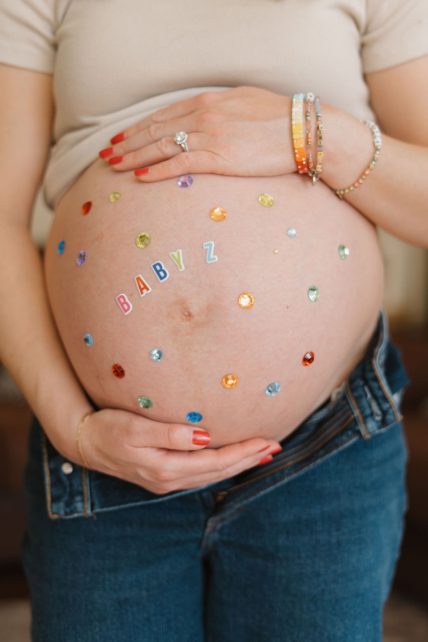 Pregnant woman's bare belly decorated with colorful gems and BABY Z letter stickers, wearing jeans and beige top.