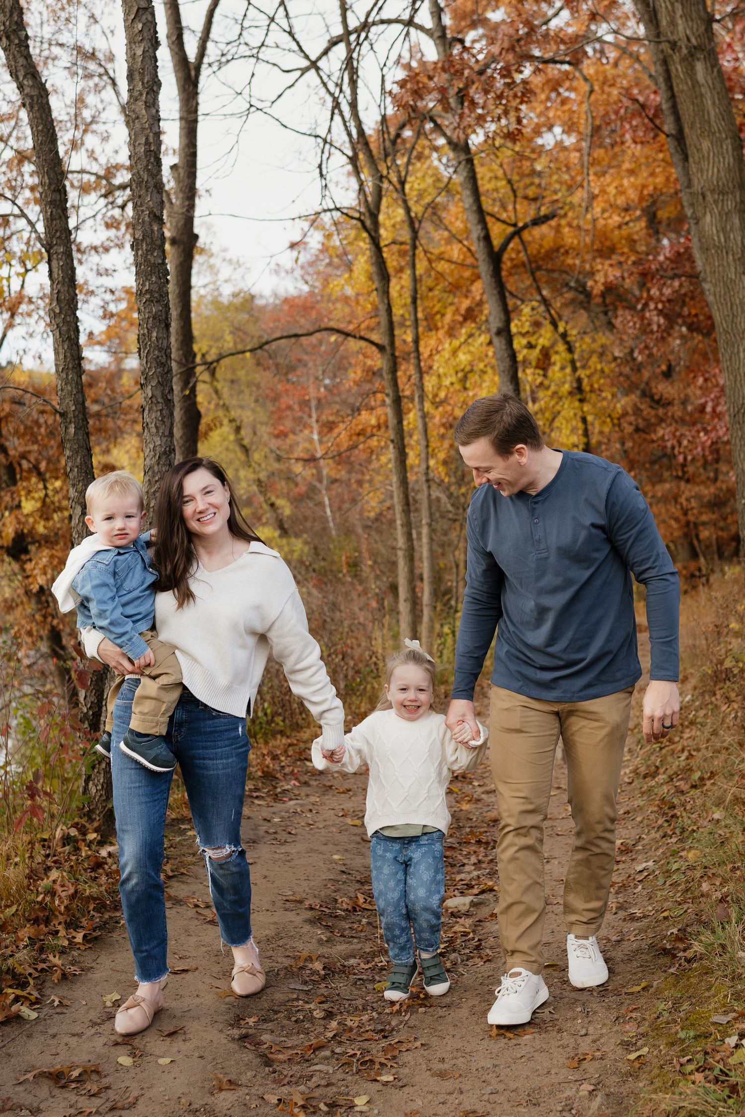 Happy family walking on autumn forest path, mother carrying baby, holding daughter's hand.