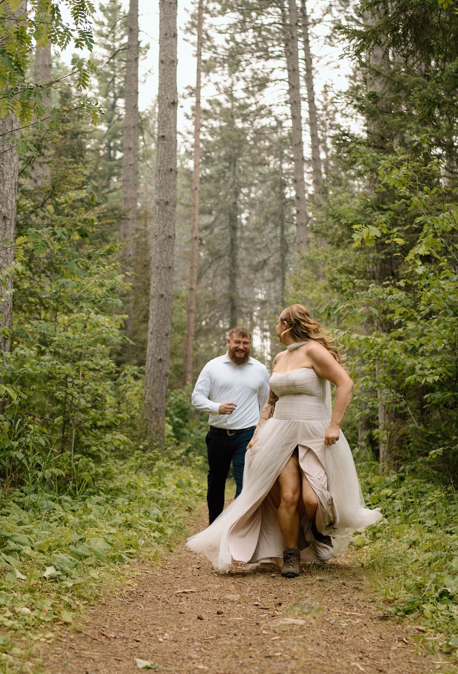 Couple walking on a forest trail, woman in elegant gown, man in casual attire, surrounded by tall pine trees.