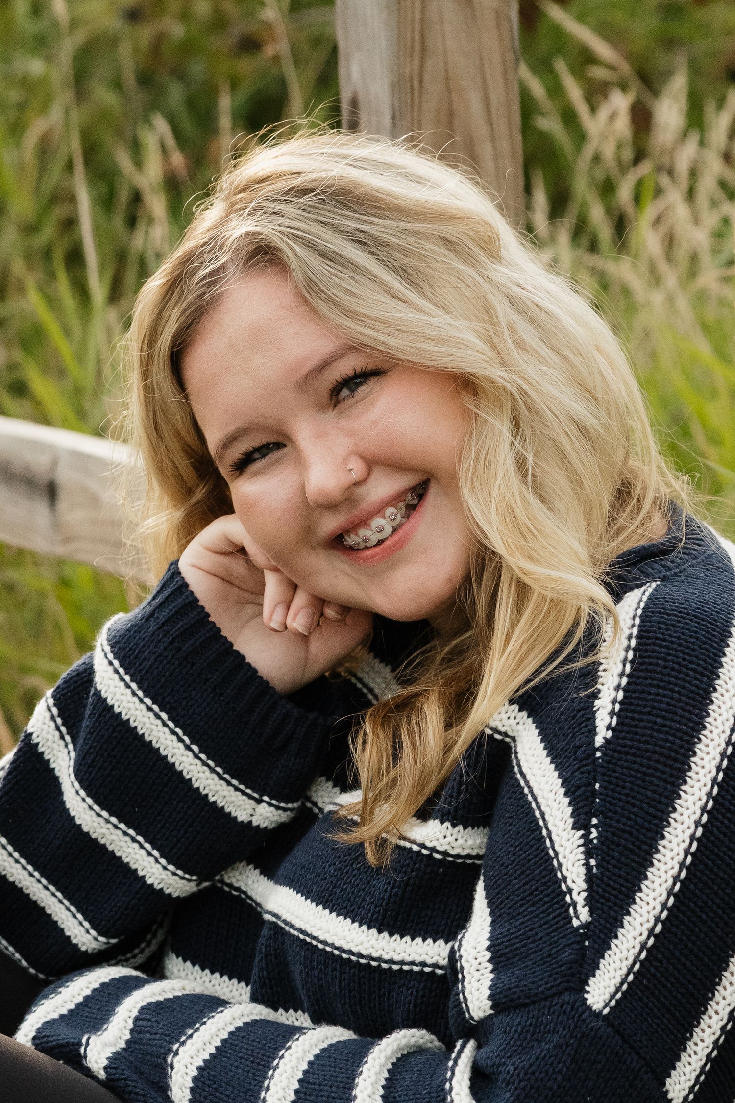 Smiling blonde teen with braces in navy striped sweater resting chin on hand near a wooden fence outdoors.