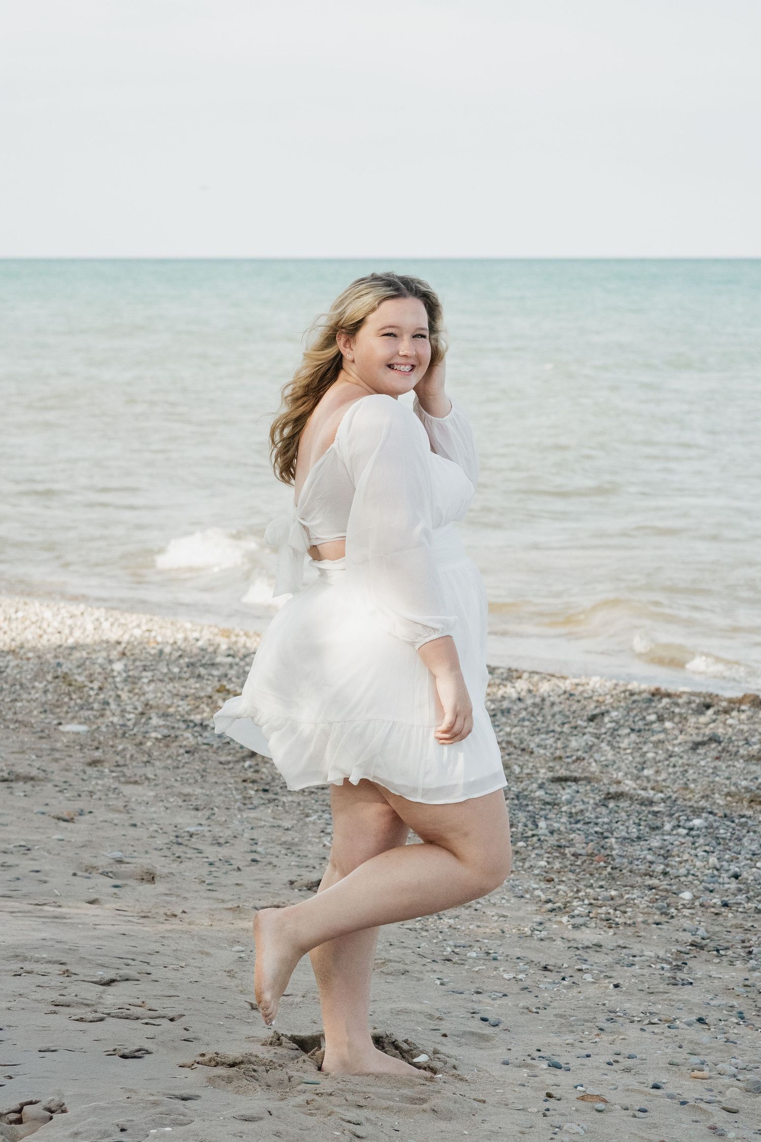Smiling blonde woman in white dress walking barefoot on pebble beach.