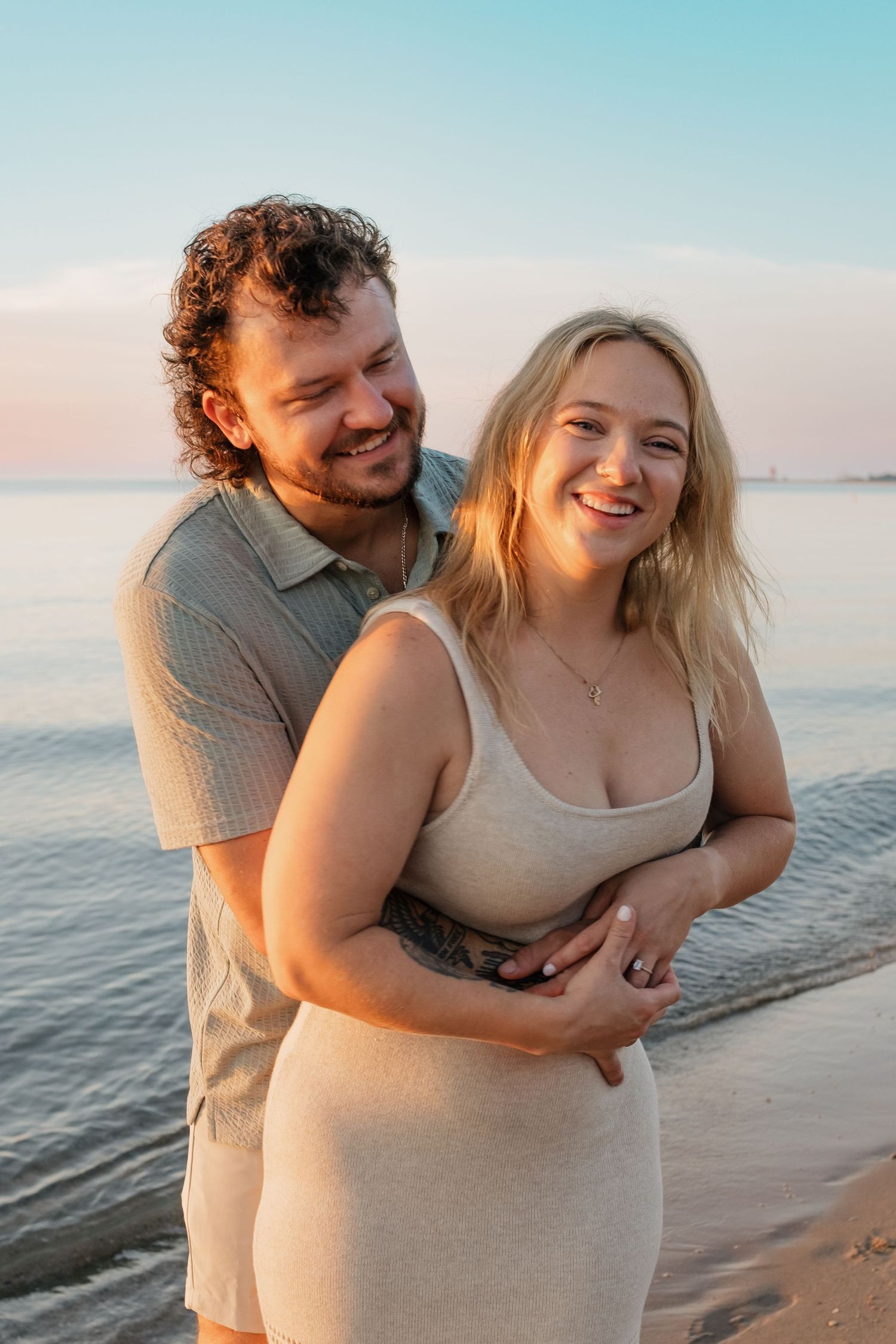 Smiling couple hugging on beach at golden hour, man wrapping arms around woman in beige fitted dress.