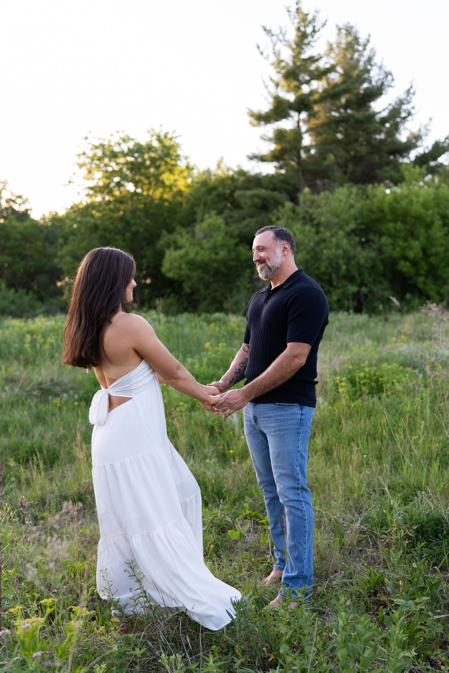 Couple holding hands in a green field at sunset, woman in white backless dress, man in black shirt and jeans.