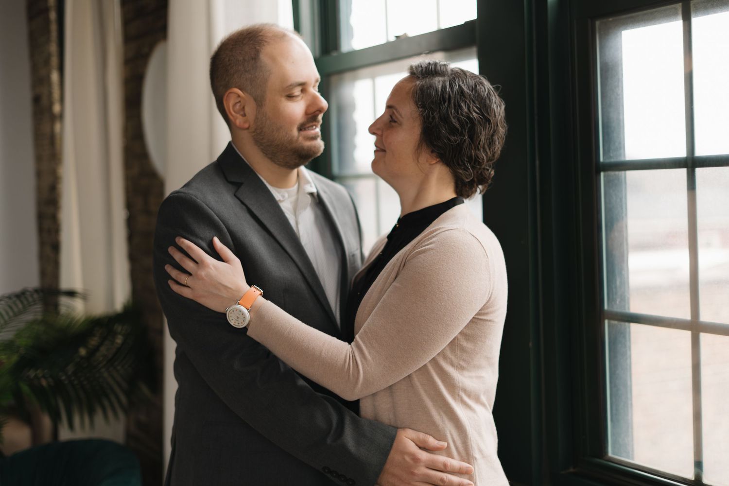 Couple embracing and gazing at each other near large windows in elegant indoor setting.