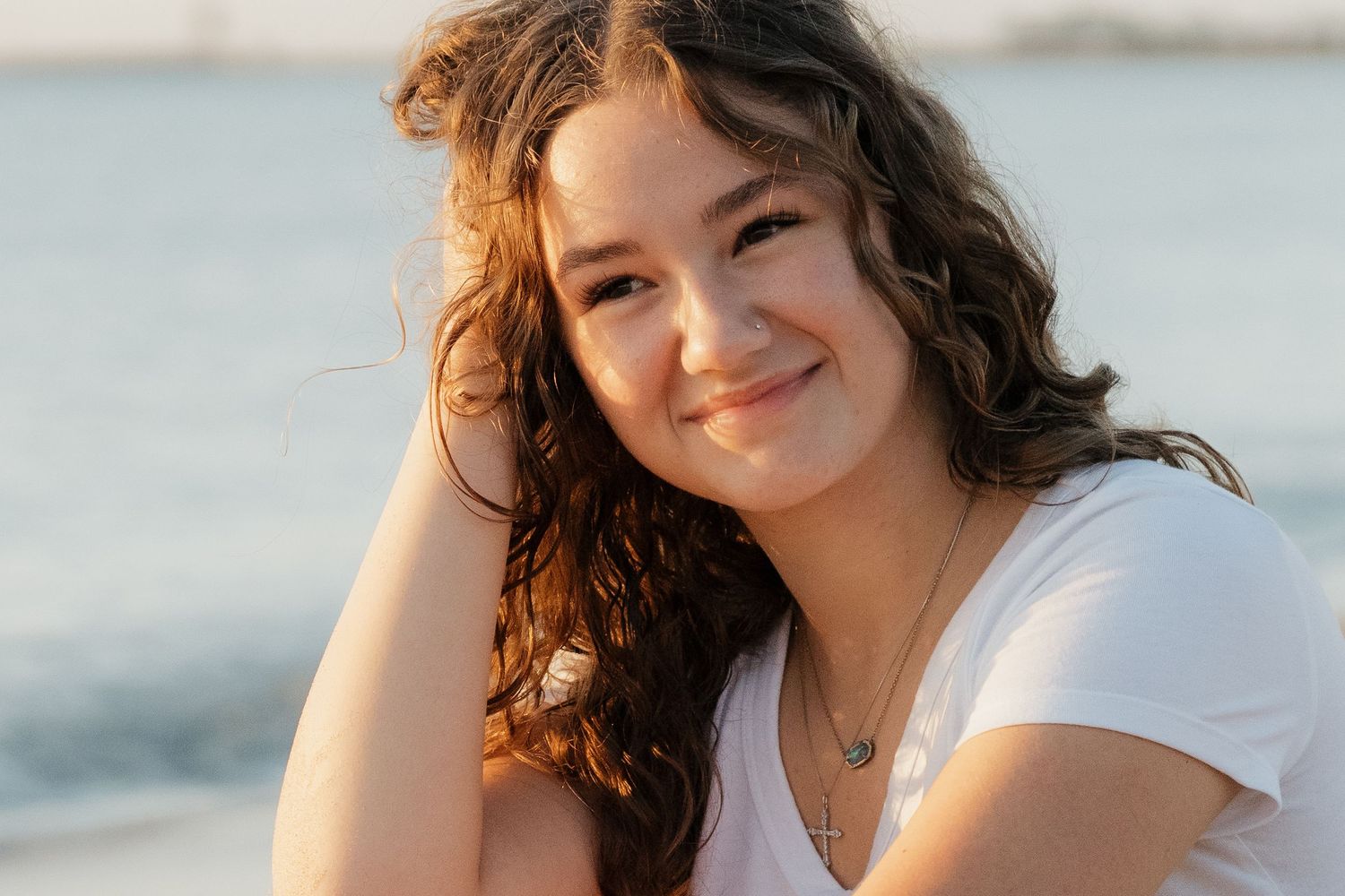 Young woman with curly hair smiling at the beach during golden hour, wearing a white t-shirt.