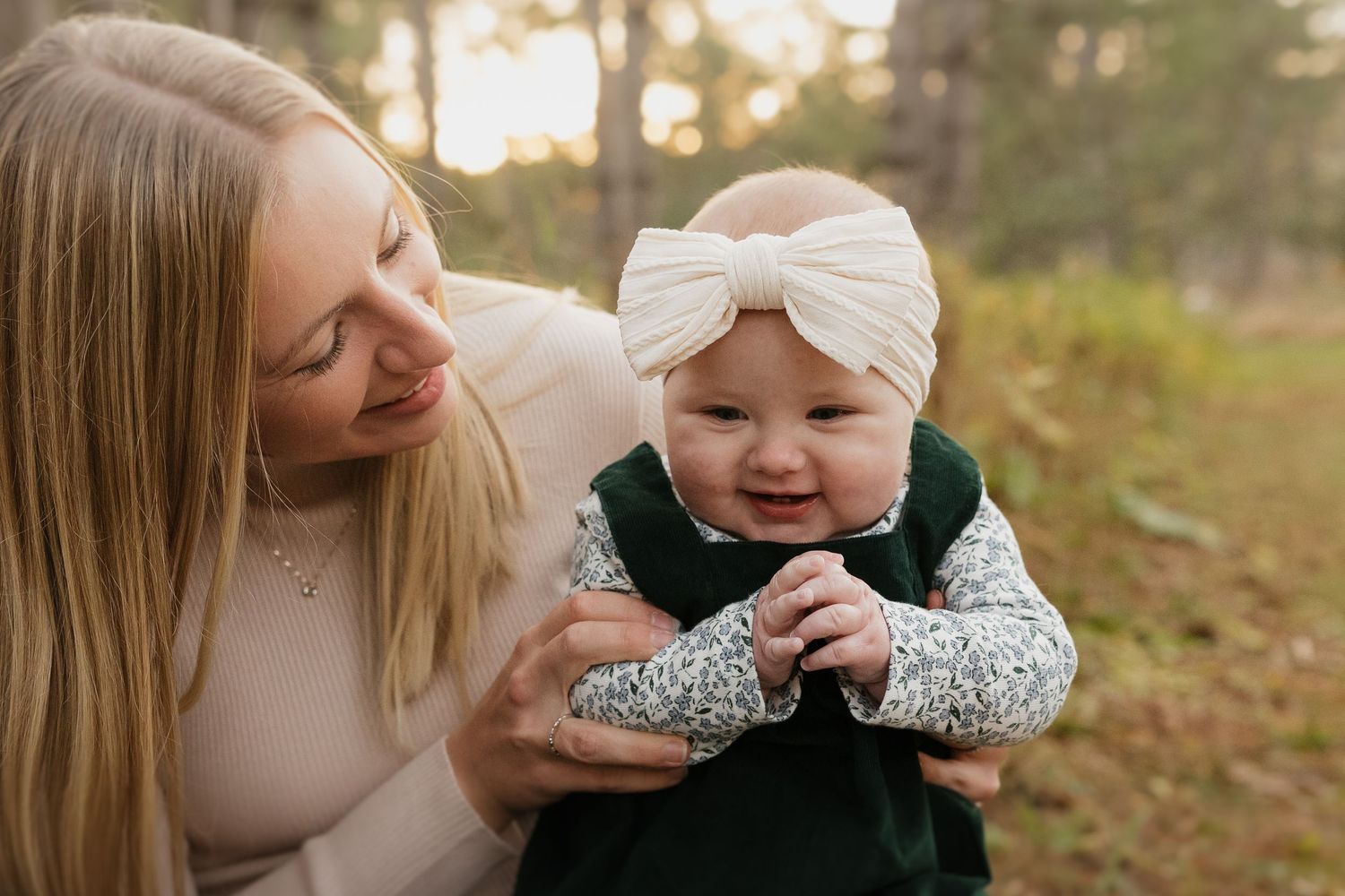 Loving mother smiling at happy baby girl wearing green outfit and large cream bow headband outdoors.
