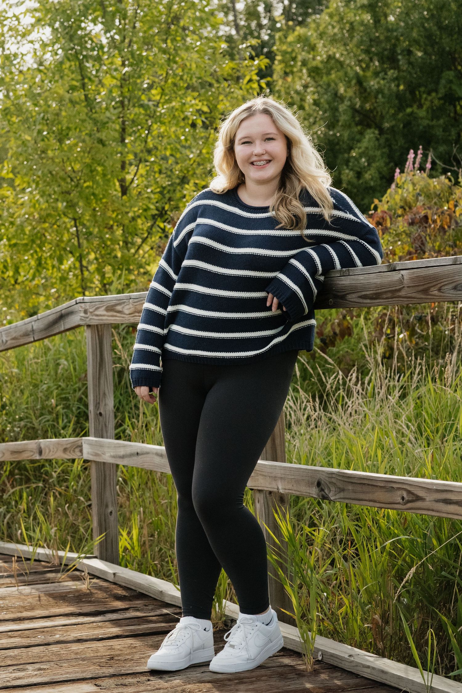 Smiling blonde teen in navy striped sweater and black leggings leaning on a wooden bridge railing outdoors.