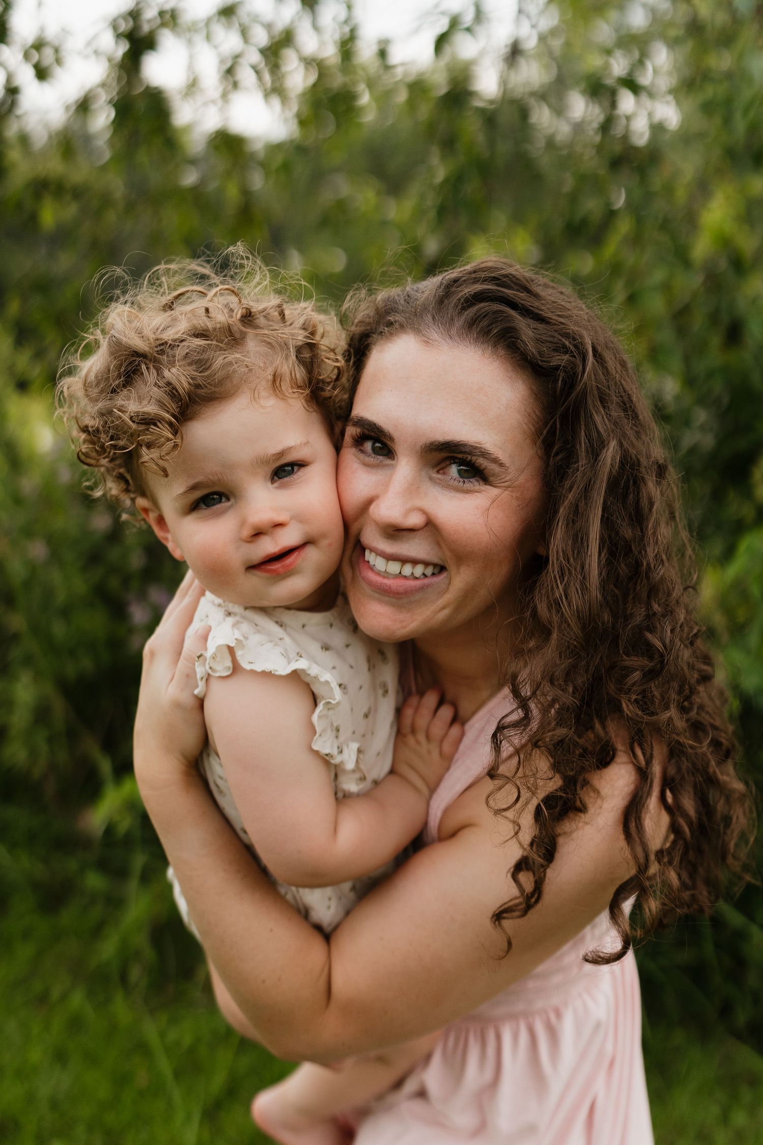 A happy mother with curly hair hugs her smiling toddler closely in a green outdoor setting.