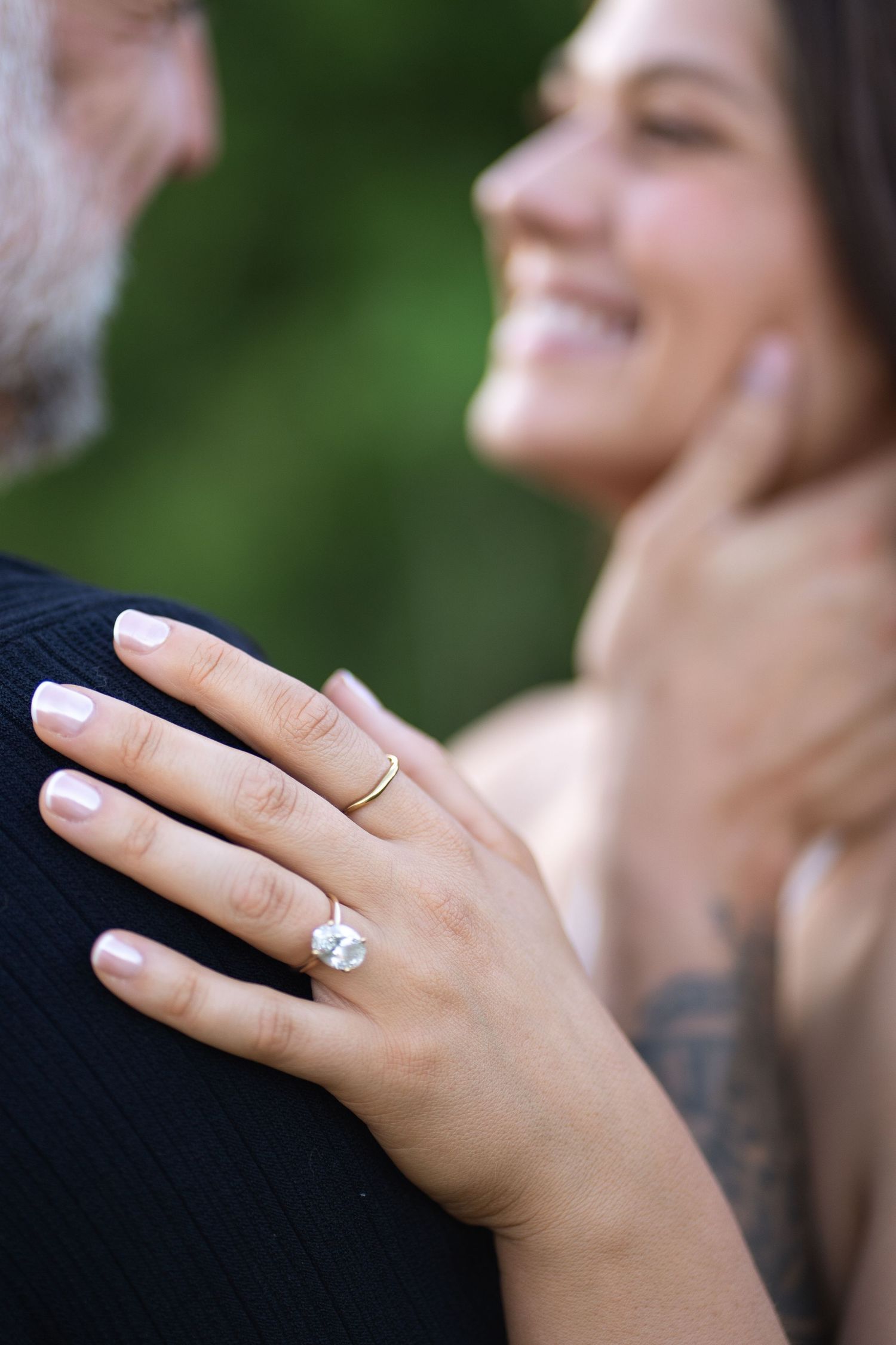 Close-up of a woman's hand with a diamond engagement ring resting on a man's shoulder, smiling couple in background.