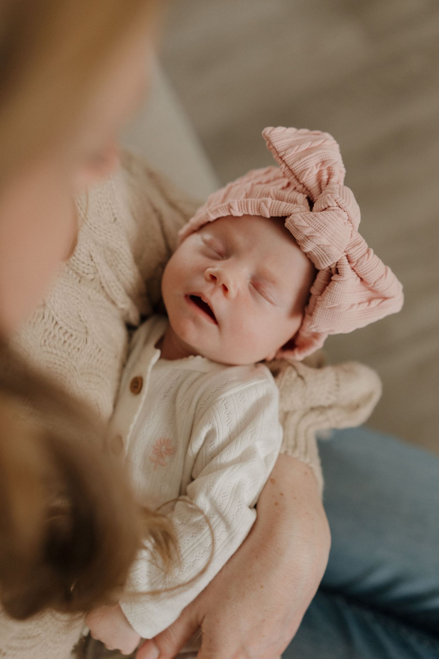 A newborn baby girl sleeping peacefully in her mother's arms, wearing a white onesie and pink bow headband.