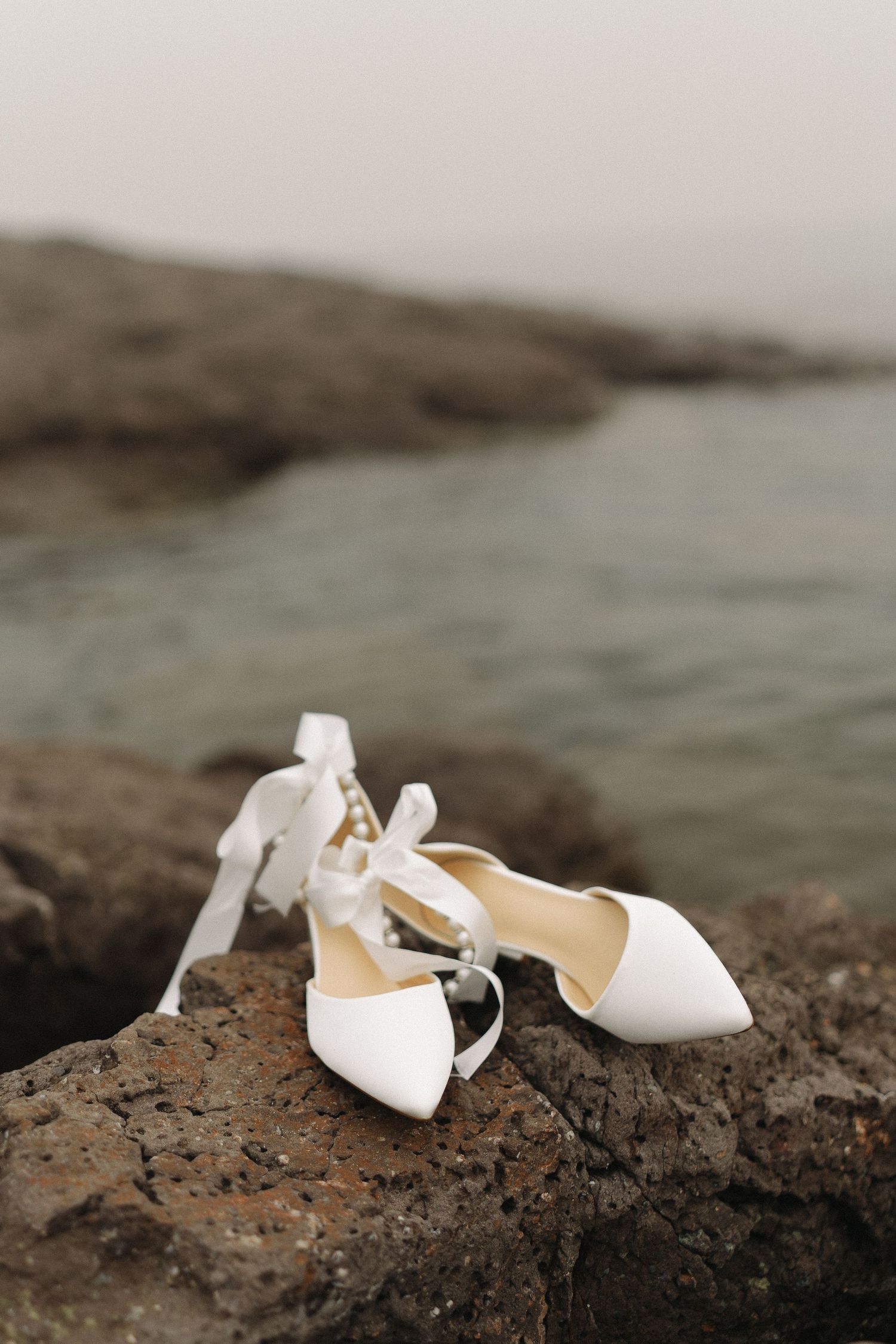 White bridal heels with ribbon ties resting on rocky shore with ocean backdrop.