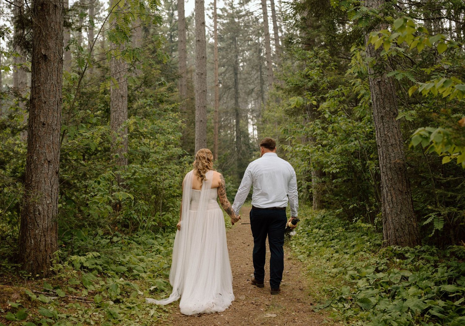 Bride and groom walking hand in hand through a lush forest on their wedding day.
