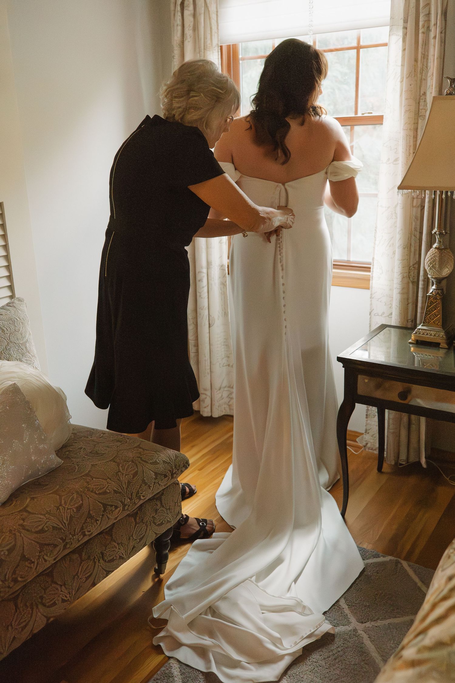 Mother helping bride button up her elegant off-shoulder white wedding gown.