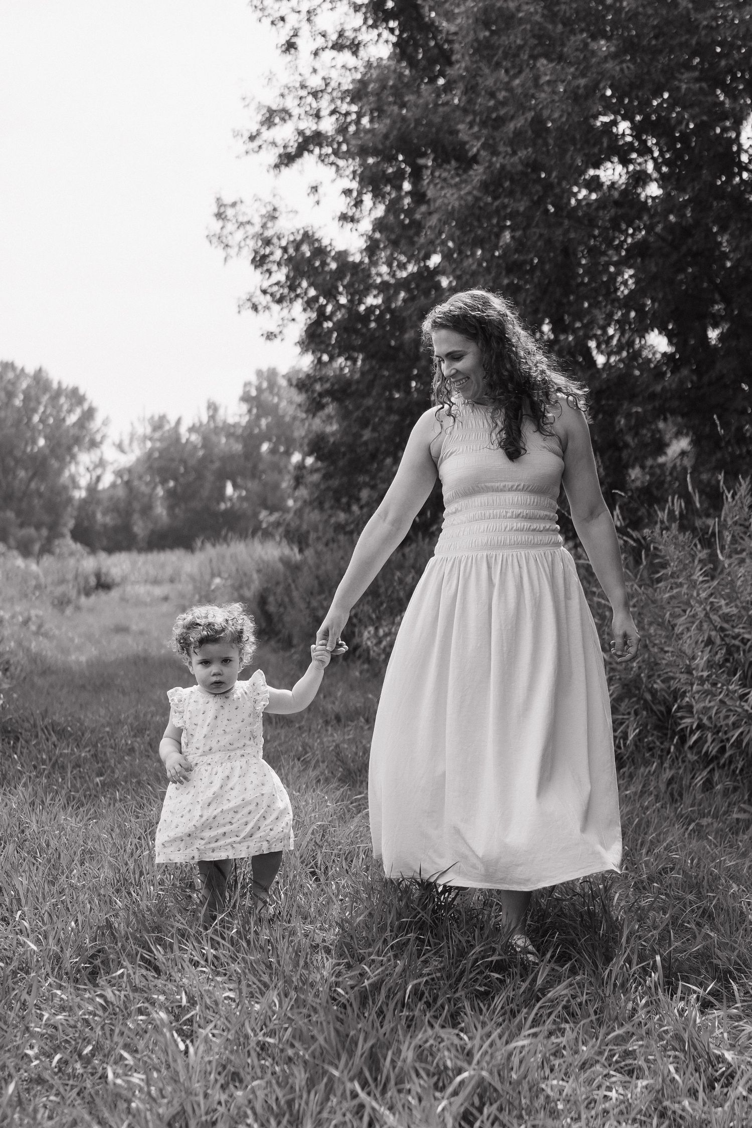 A black and white photo of a mother and toddler holding hands while walking through a field.