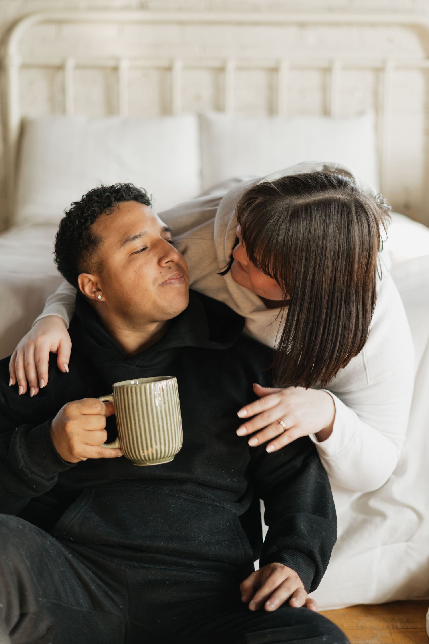 A couple sharing a tender moment, woman kissing man's cheek as he holds a ceramic mug, sitting by a bed.