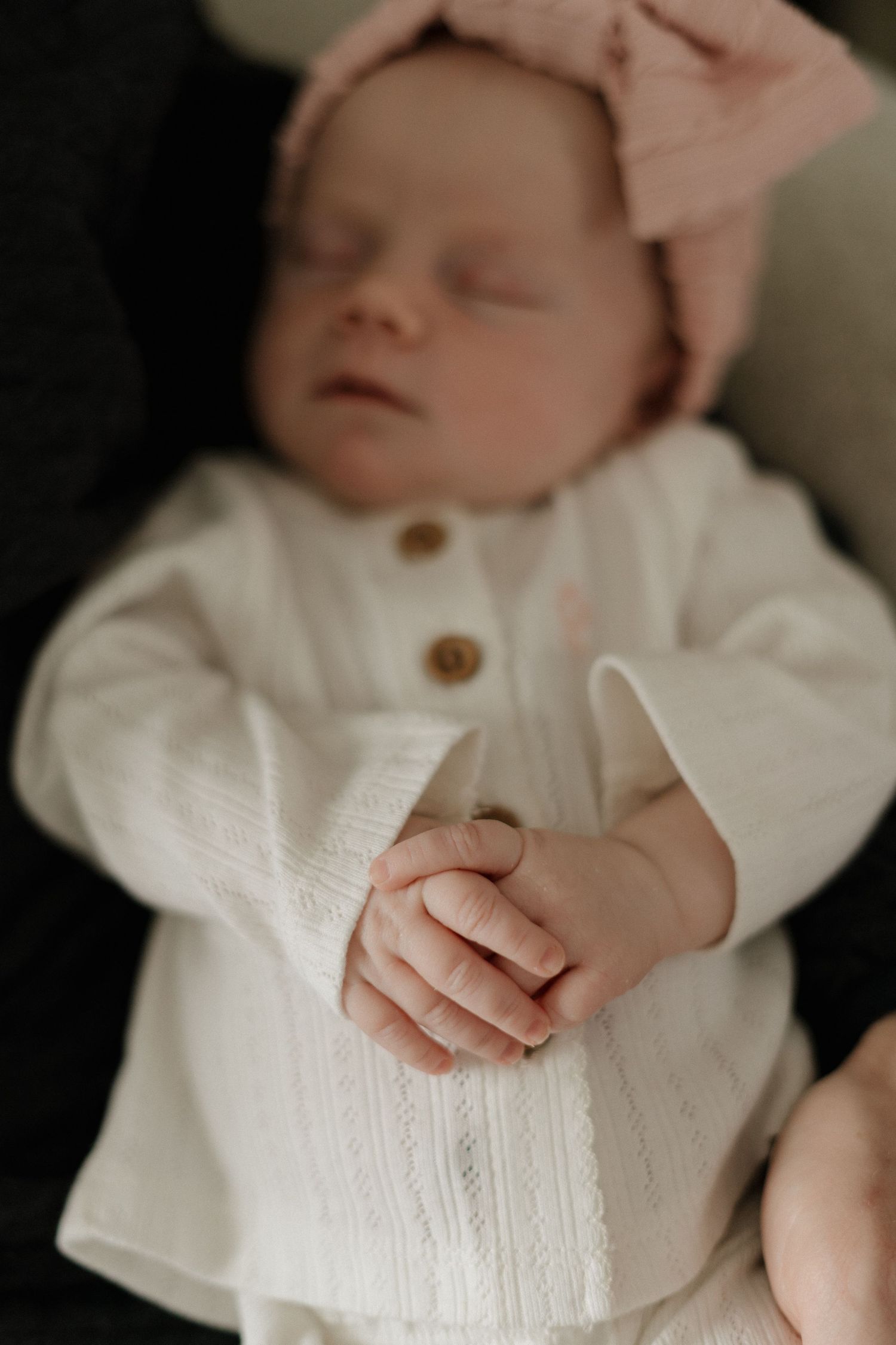 Sleeping newborn baby in white knit outfit and pink bow headband with tiny hands clasped together.
