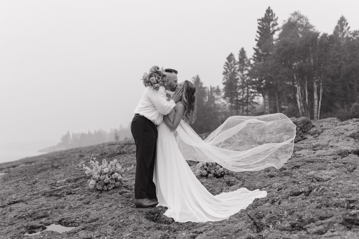Black and white photo of a bride and groom embracing outdoors, her veil flowing in the wind near a rocky shoreline.