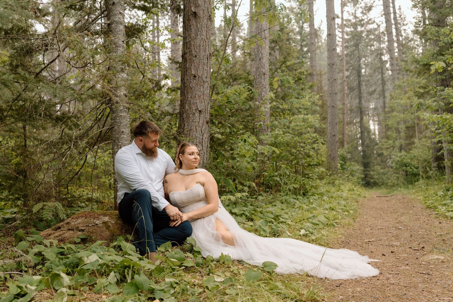 Bride in white gown and groom in jeans sit together on a forest path surrounded by tall pine trees.
