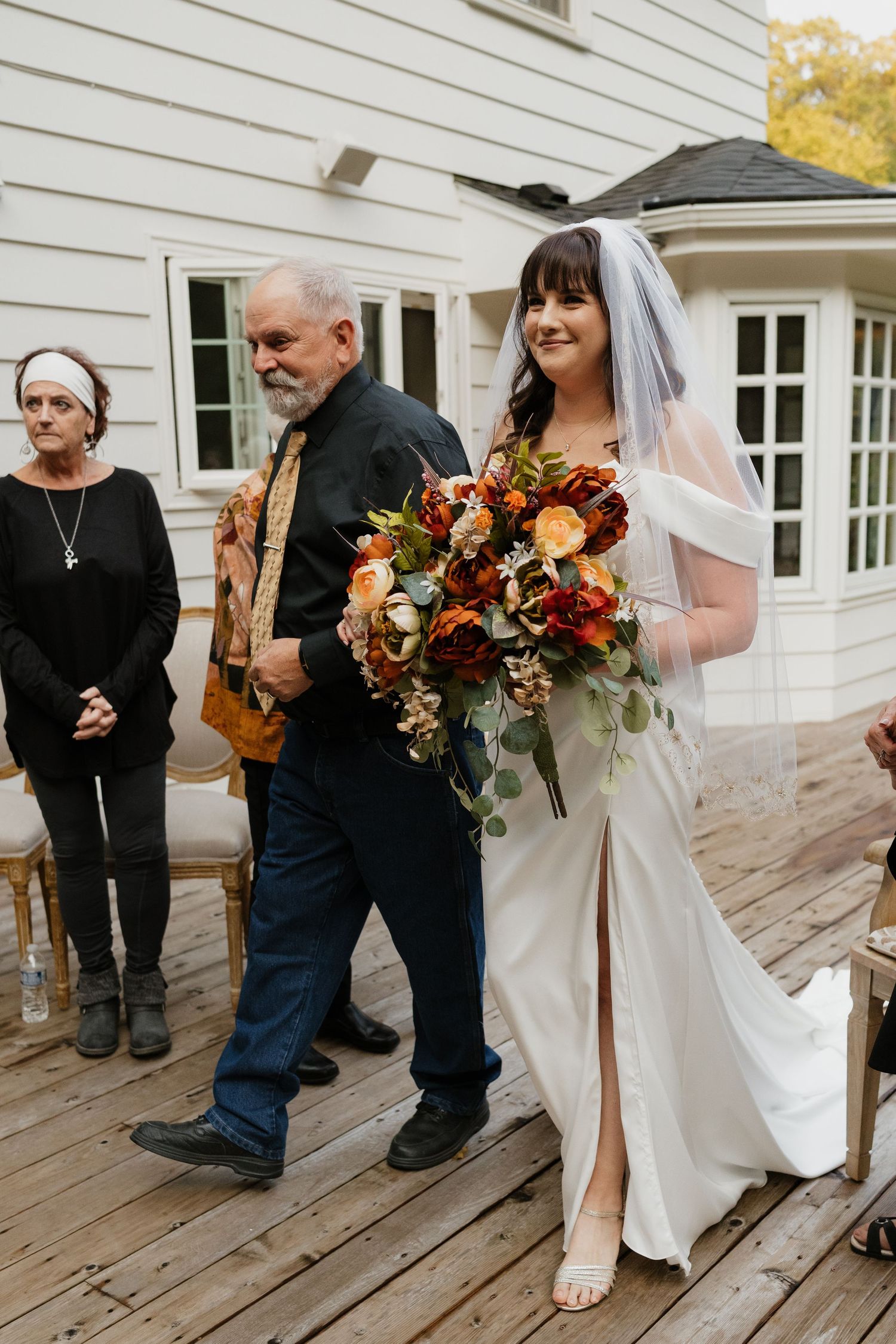 Smiling bride in white gown holding autumn bouquet walks down aisle with her father.