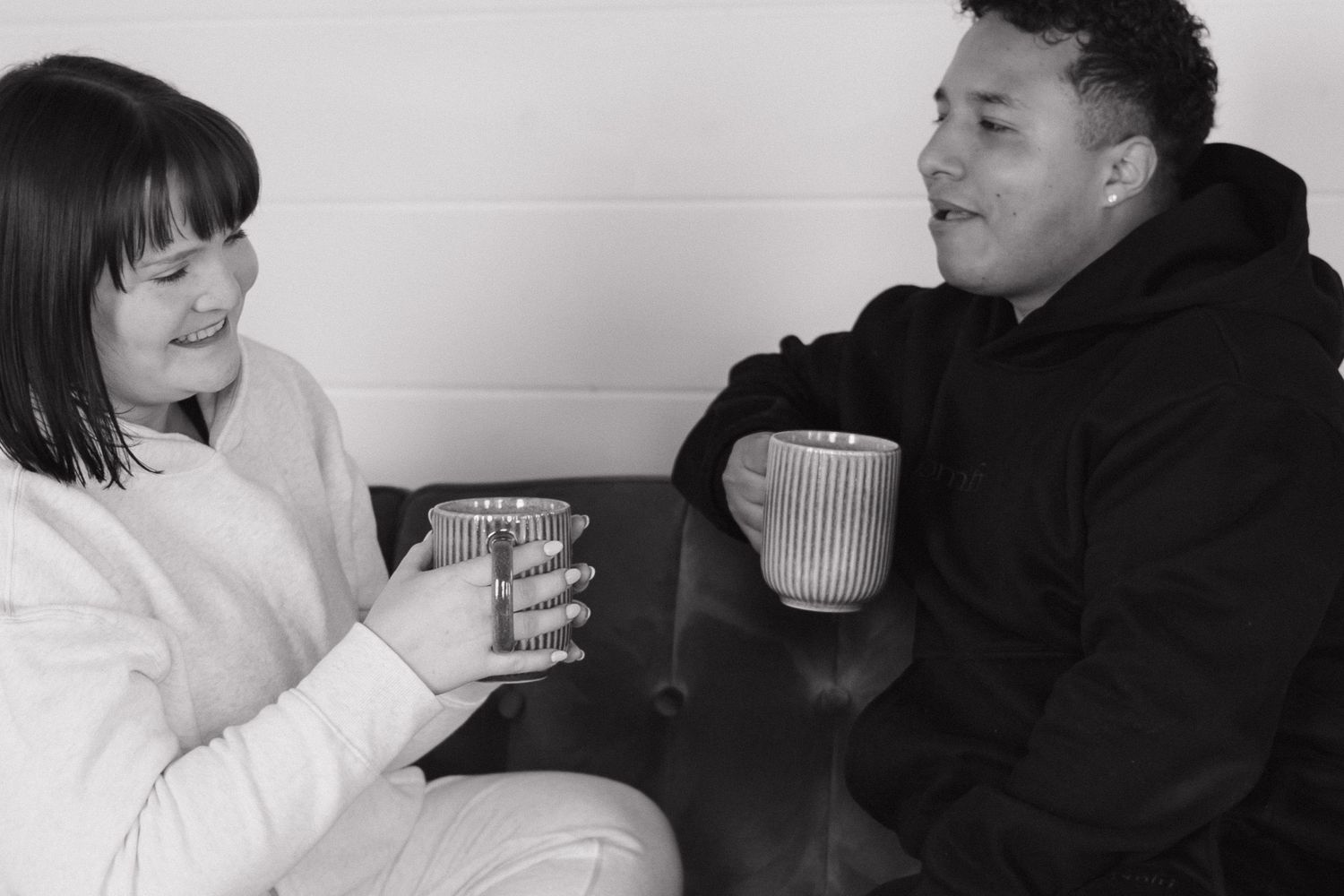 Two people sitting together smiling and clinking mugs in a cozy black and white photo.