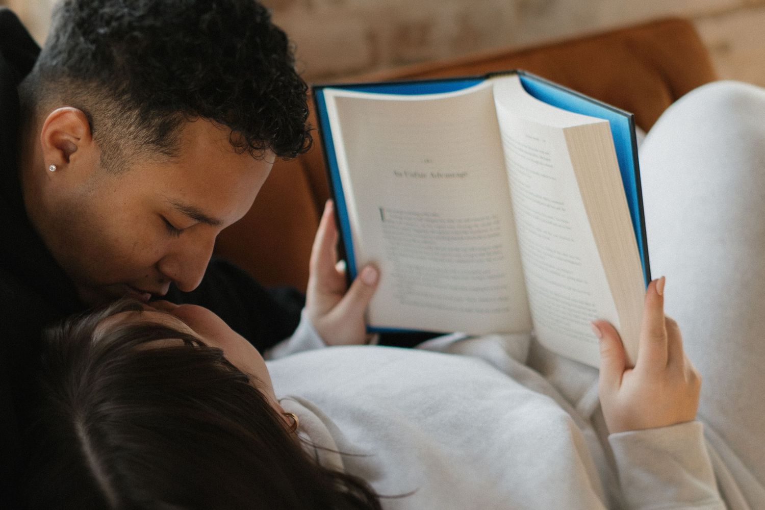 Couple relaxing together on a couch, man kissing woman's head while she reads an open book.