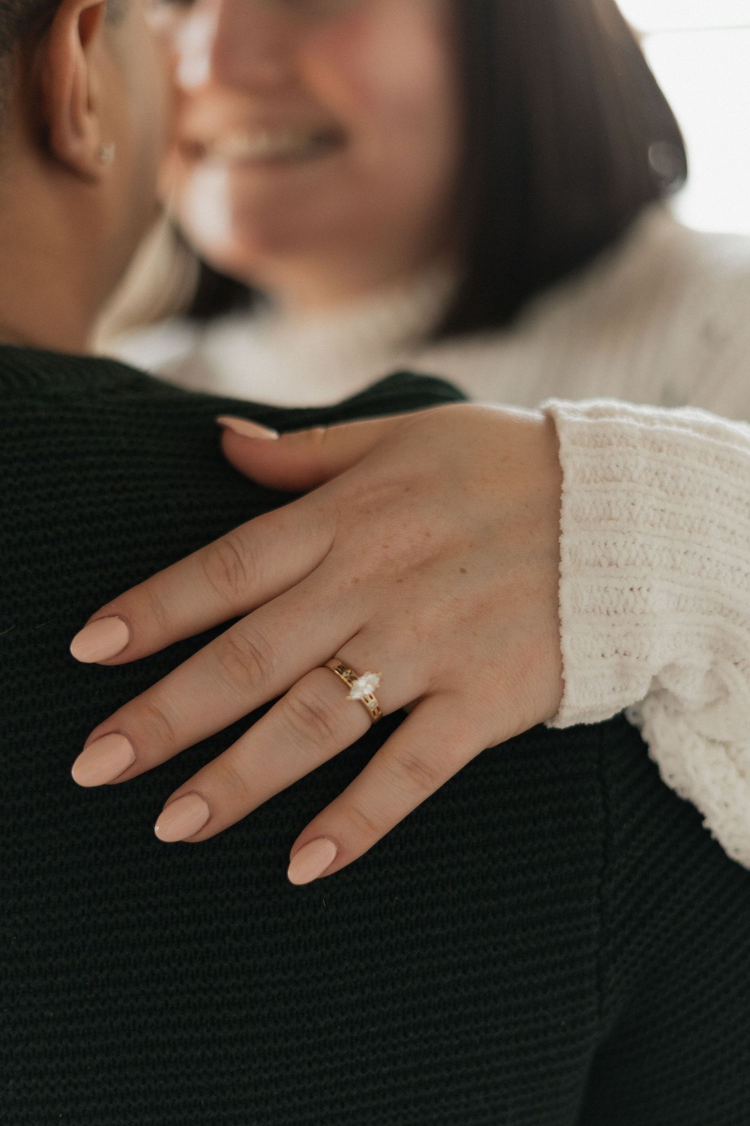 Close-up of a woman's hand with nude nails wearing a gold solitaire engagement ring, resting on a man's shoulder.