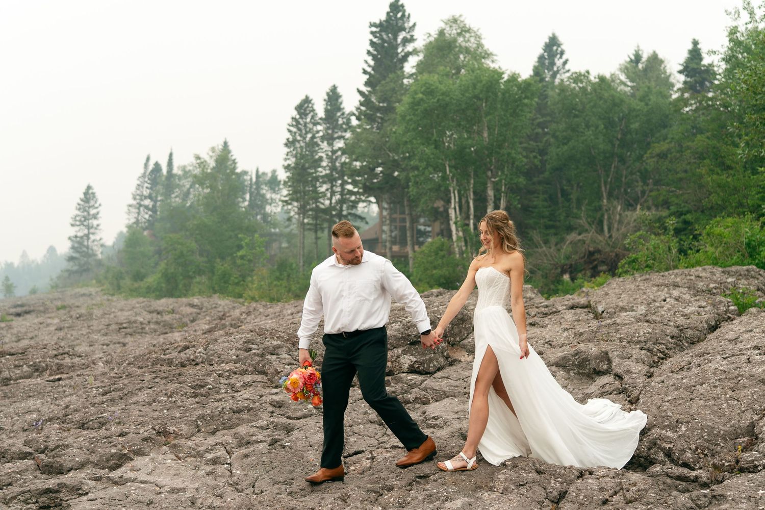 Bride and groom hold hands walking on rocky shoreline with misty forest backdrop during outdoor wedding.