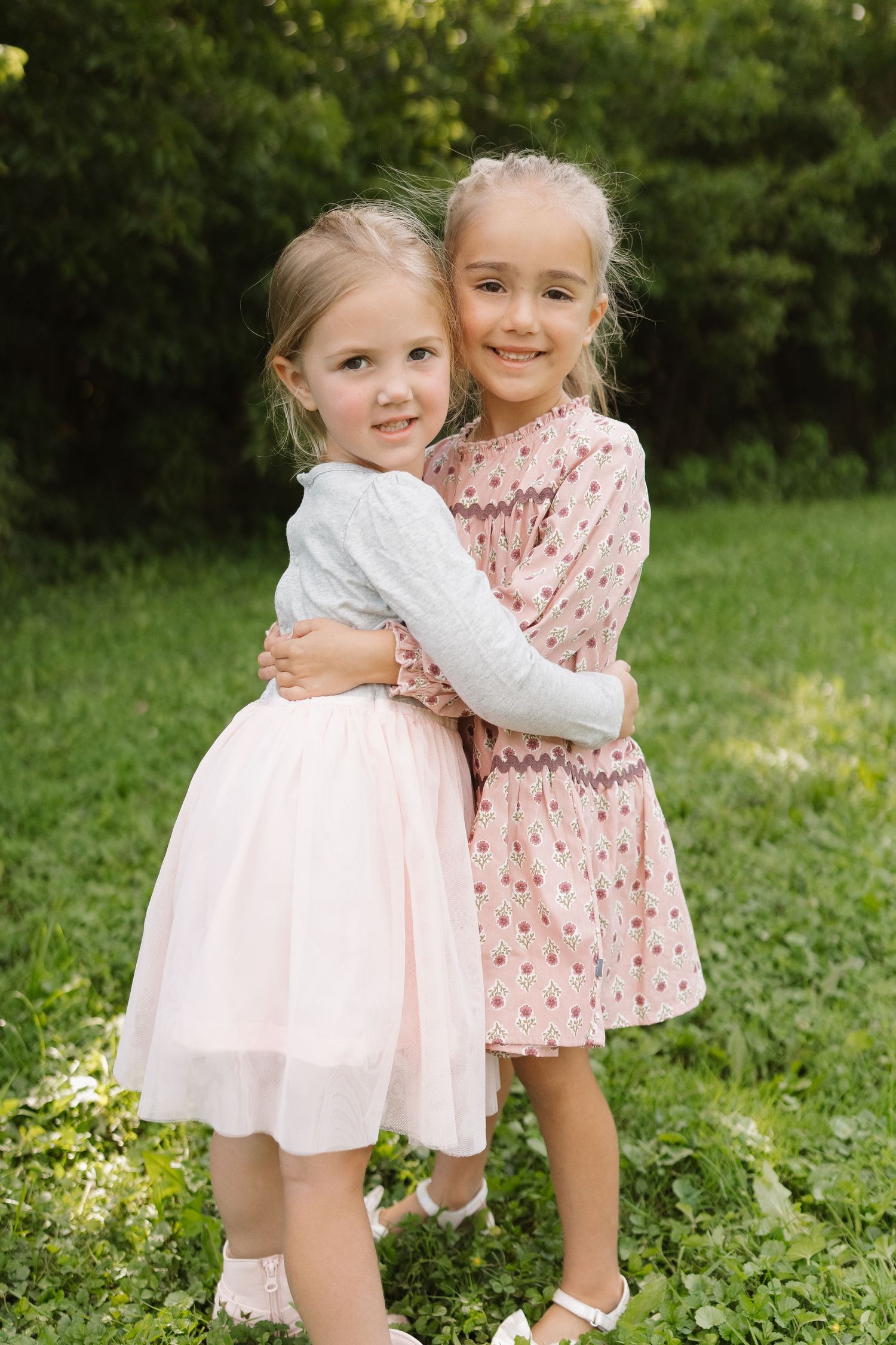 Two young girls in pretty dresses hug each other while standing on green grass outdoors.