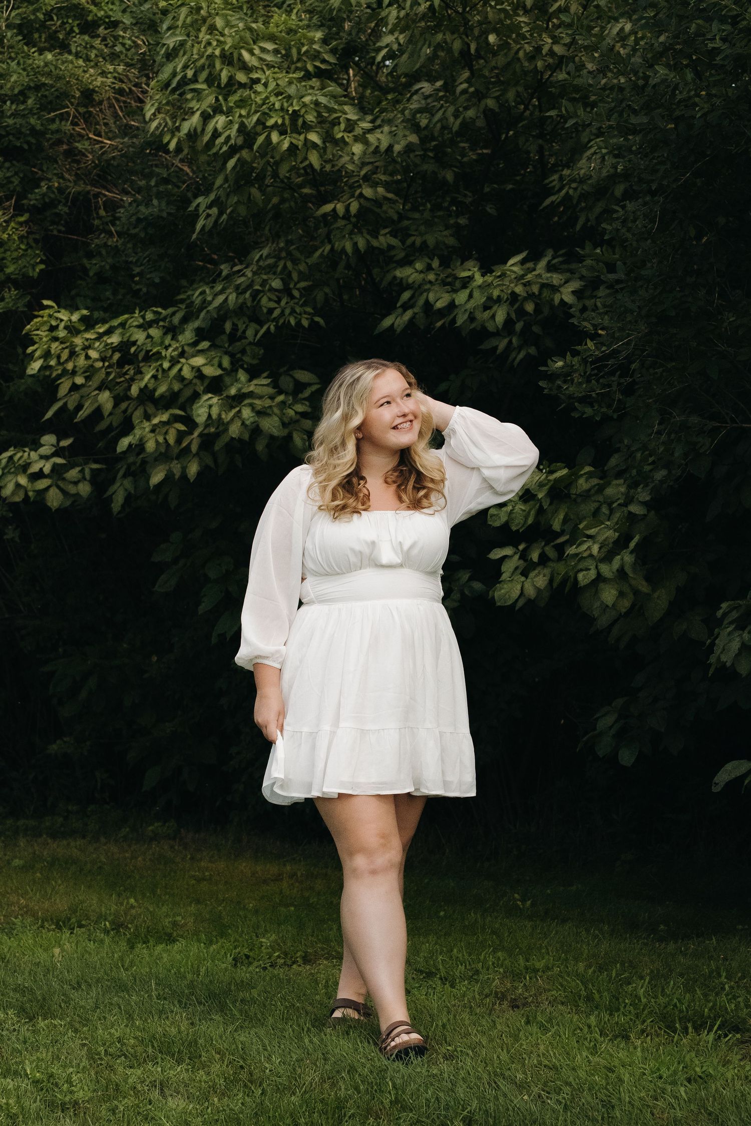 Young blonde woman in white mini dress poses on green grass with lush trees in background.