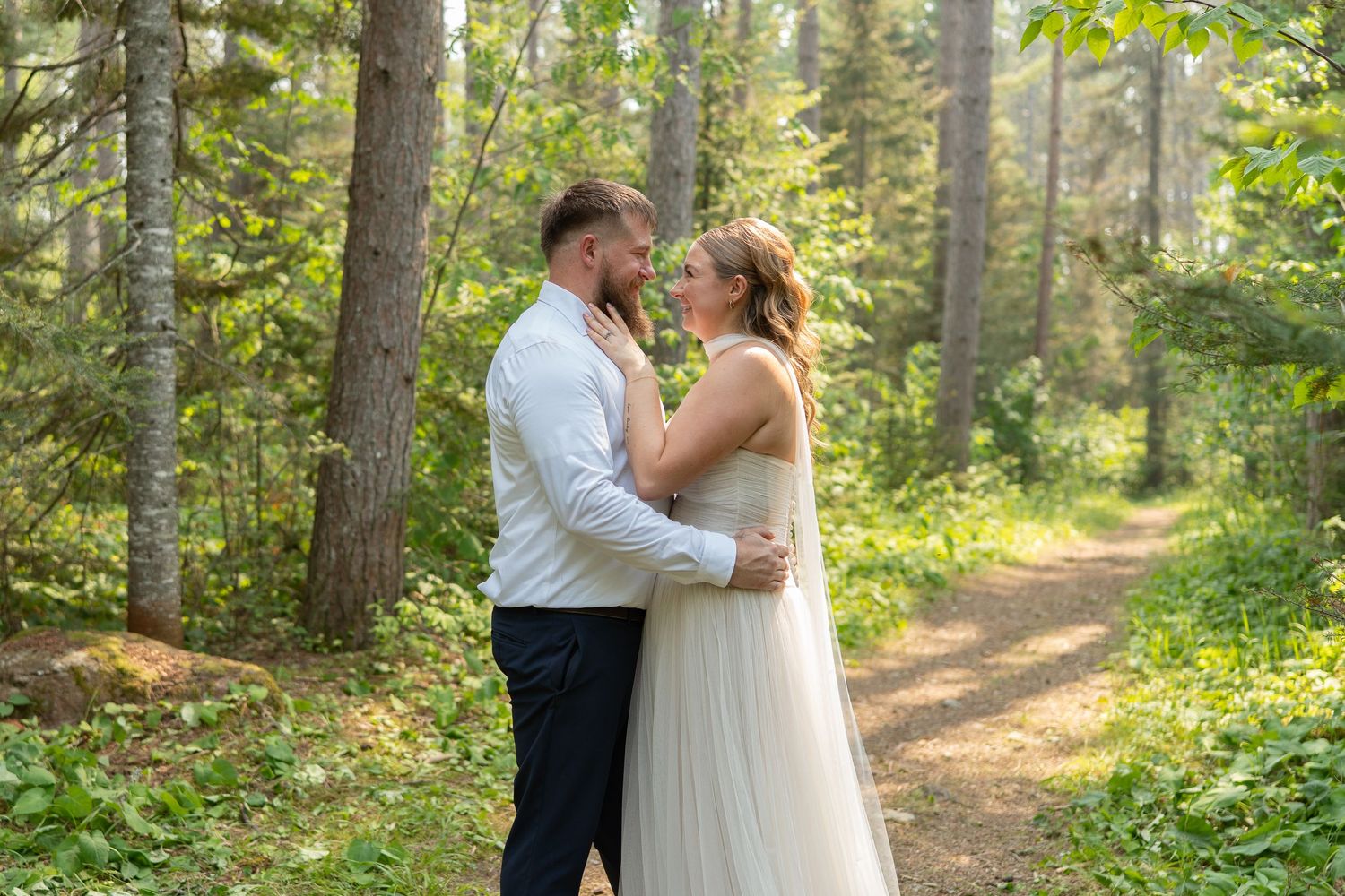 A bride and groom share a romantic embrace on a forest path surrounded by lush green trees on their wedding day.