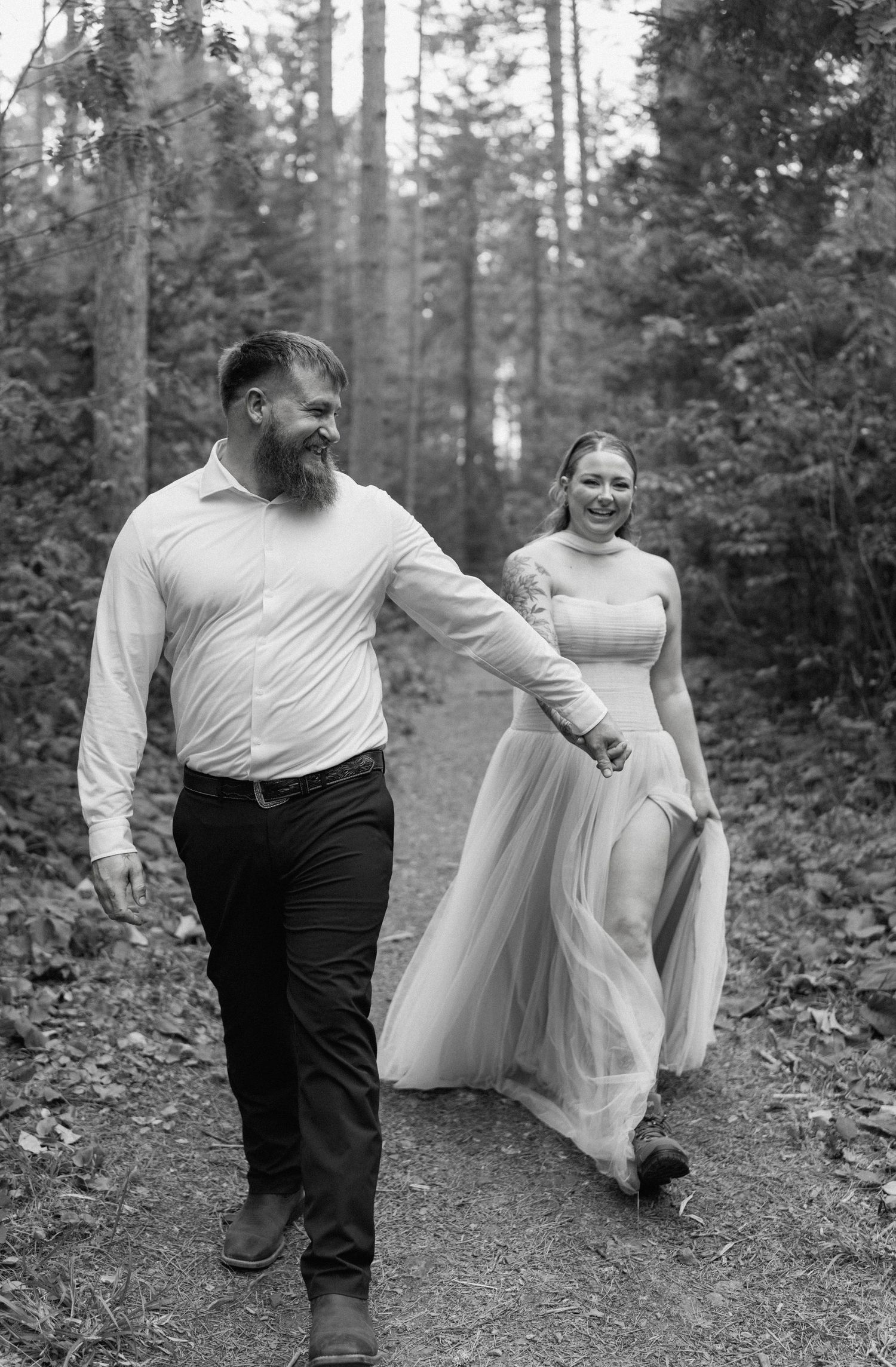 Black and white photo of a bearded groom leading his smiling bride by the hand along a forest path.