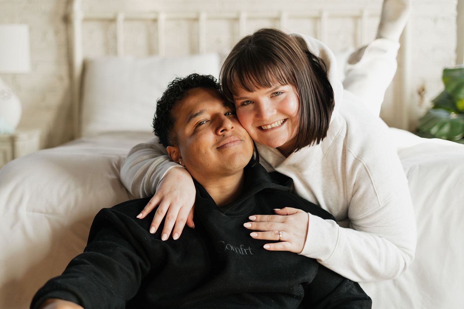 A smiling interracial couple cuddles together on a bed, woman in white sweater embracing man in black hoodie.