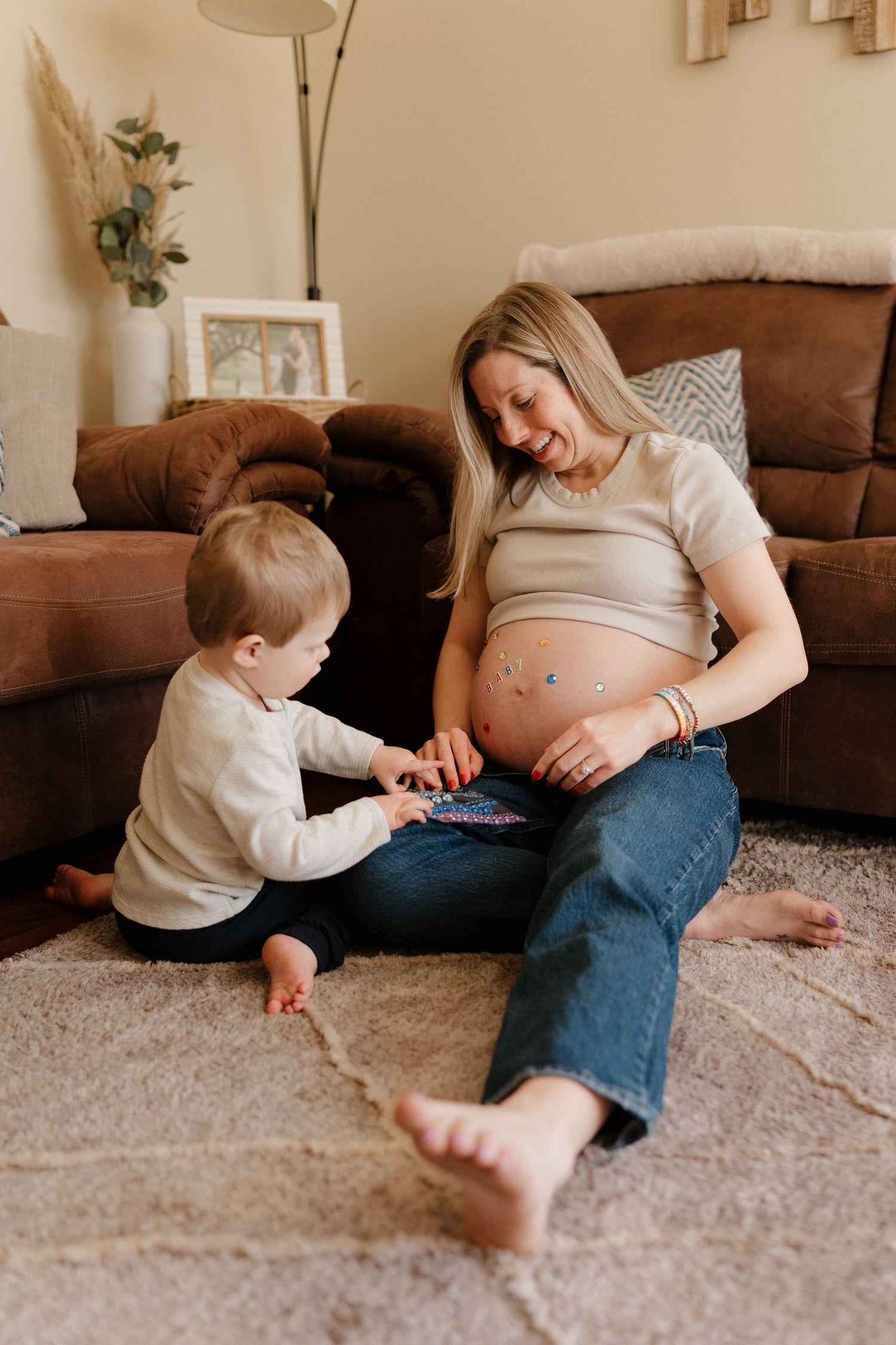 Pregnant woman sitting on floor with toddler placing stickers on her baby bump in a cozy living room.