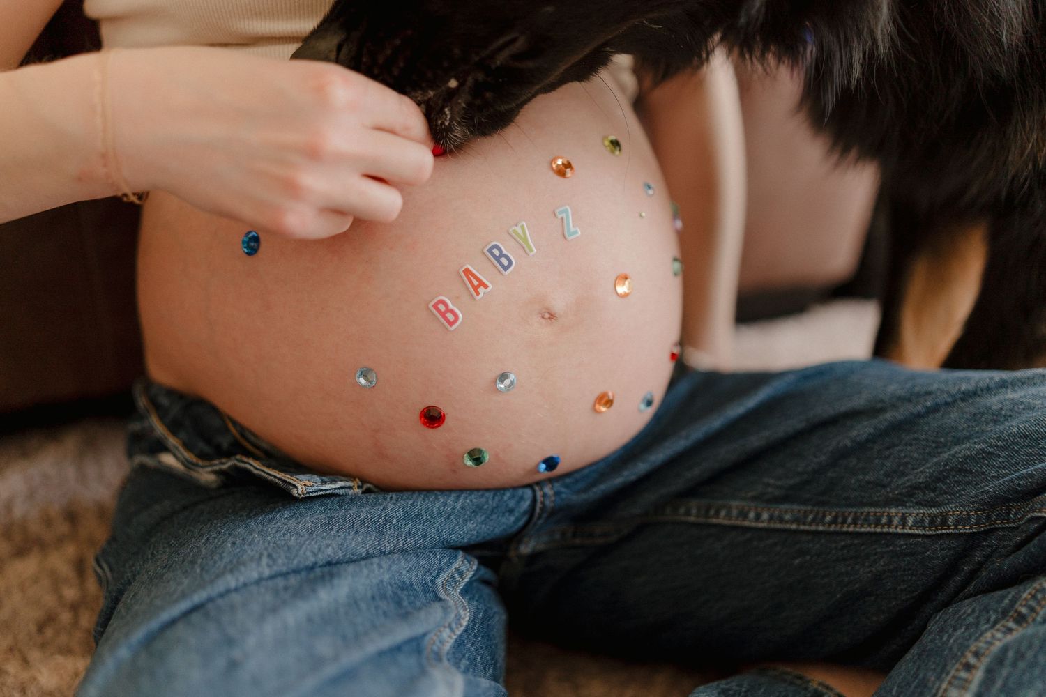 Pregnant woman sitting with colorful letter stickers spelling BABY on her bare baby bump, wearing blue jeans.