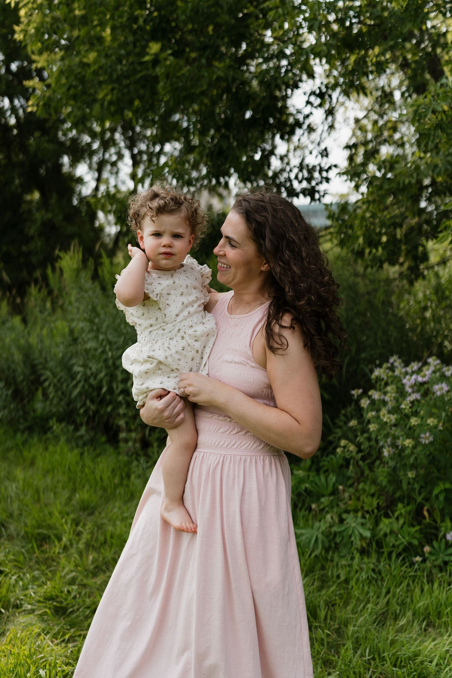 A smiling mother in a pink dress holds her curly-haired toddler among lush green trees.