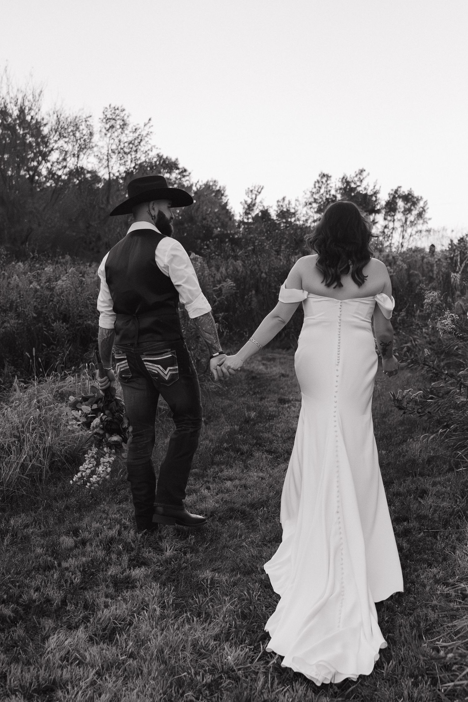 Black and white photo of bride and groom holding hands walking through a field on their wedding day.