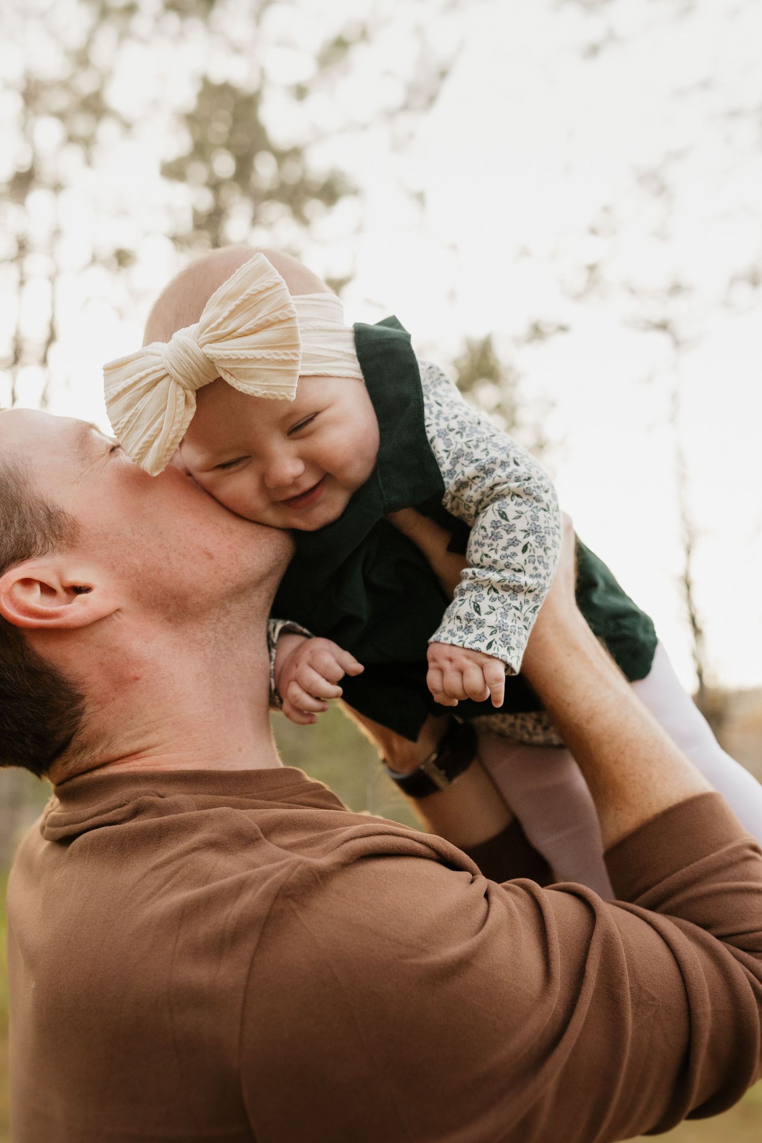Father kissing smiling baby girl with cream bow headband while lifting her up in pine forest.