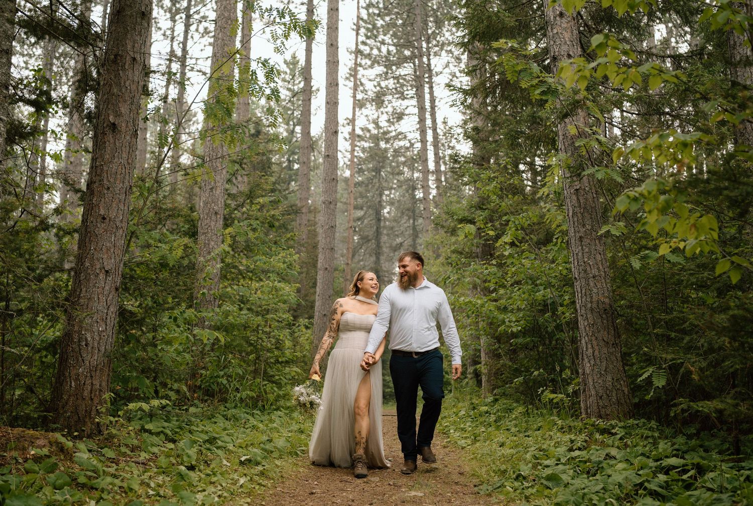 Couple walks hand-in-hand through lush forest on wedding day, bride in flowing gown, groom in white shirt.